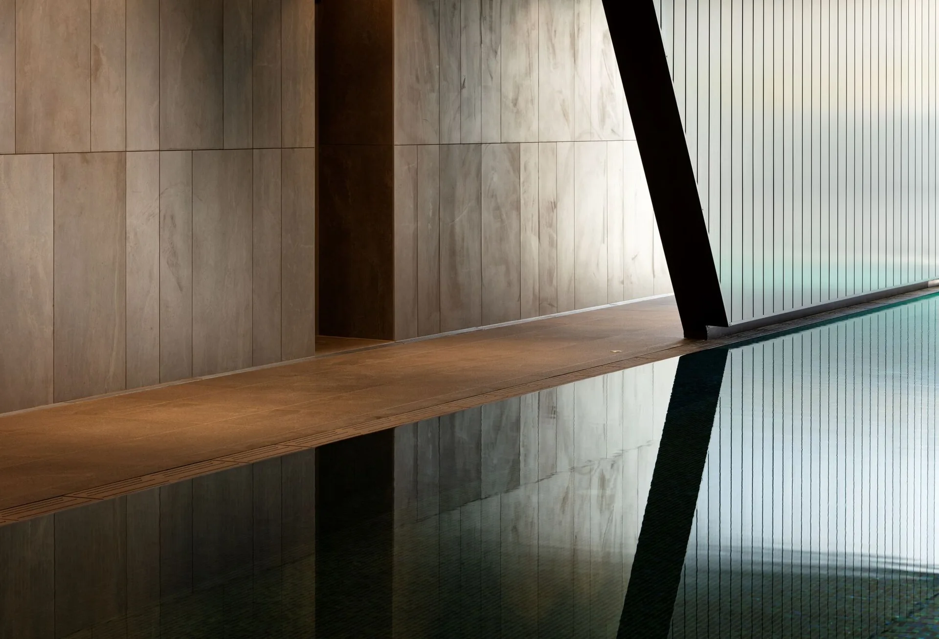 An indoor swimming pool with reflective water next to a beige stone tile walkway, a vertically paneled wall, and a slatted glass partition.