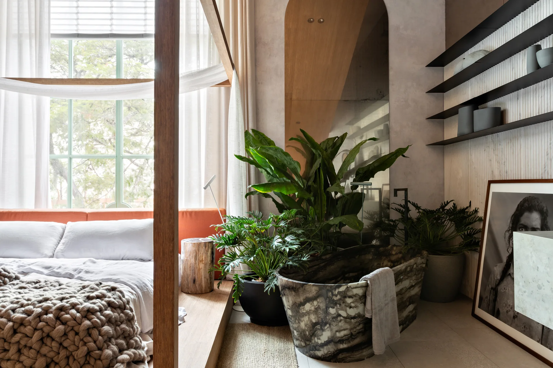 A bedroom suite features a wood platform canopy bed, an organic stone-like freestanding bathtub, and lush potted green plants under a large mirror.