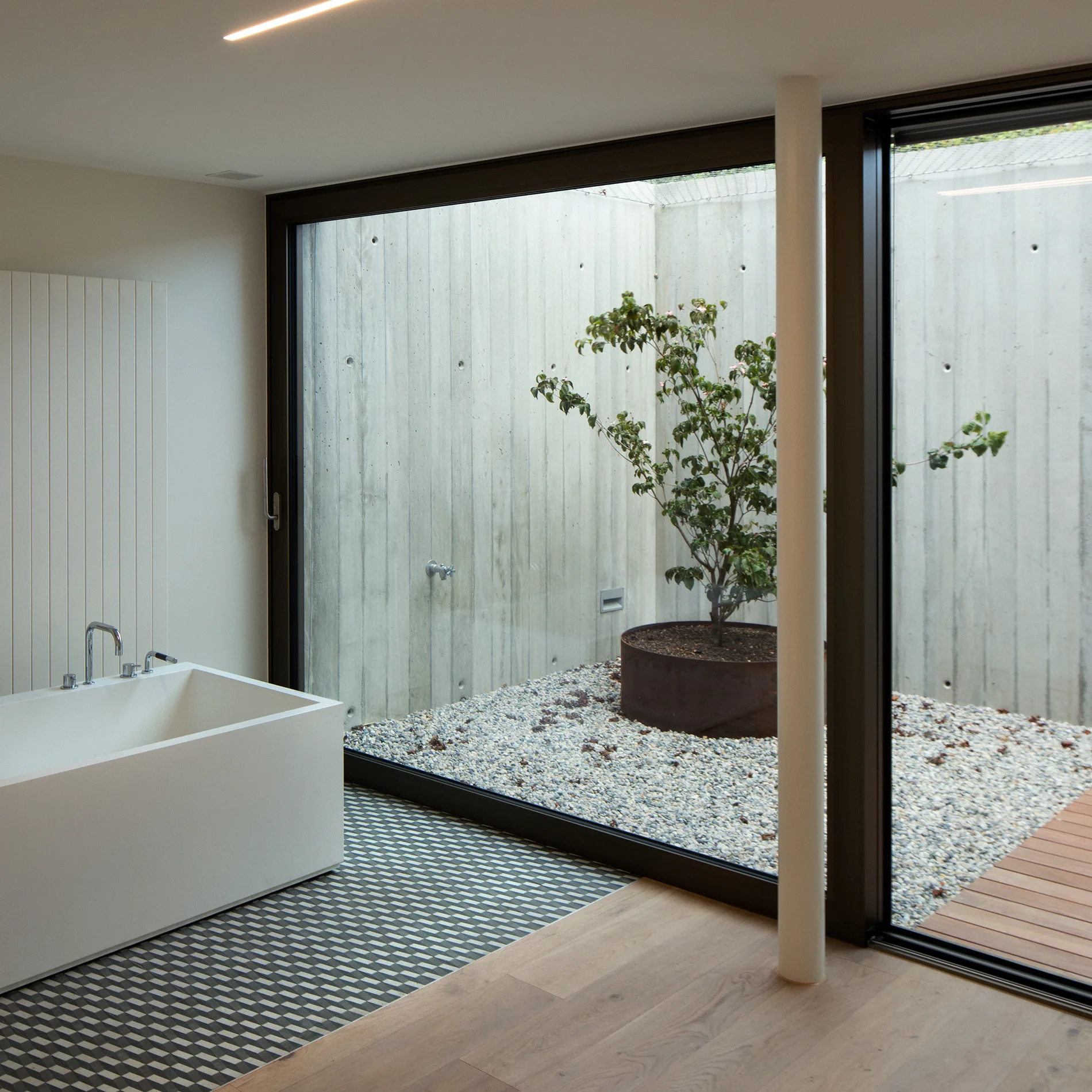 A modern bathroom features a white rectangular bathtub on geometric tile next to glass walls overlooking an enclosed concrete courtyard with a tree.