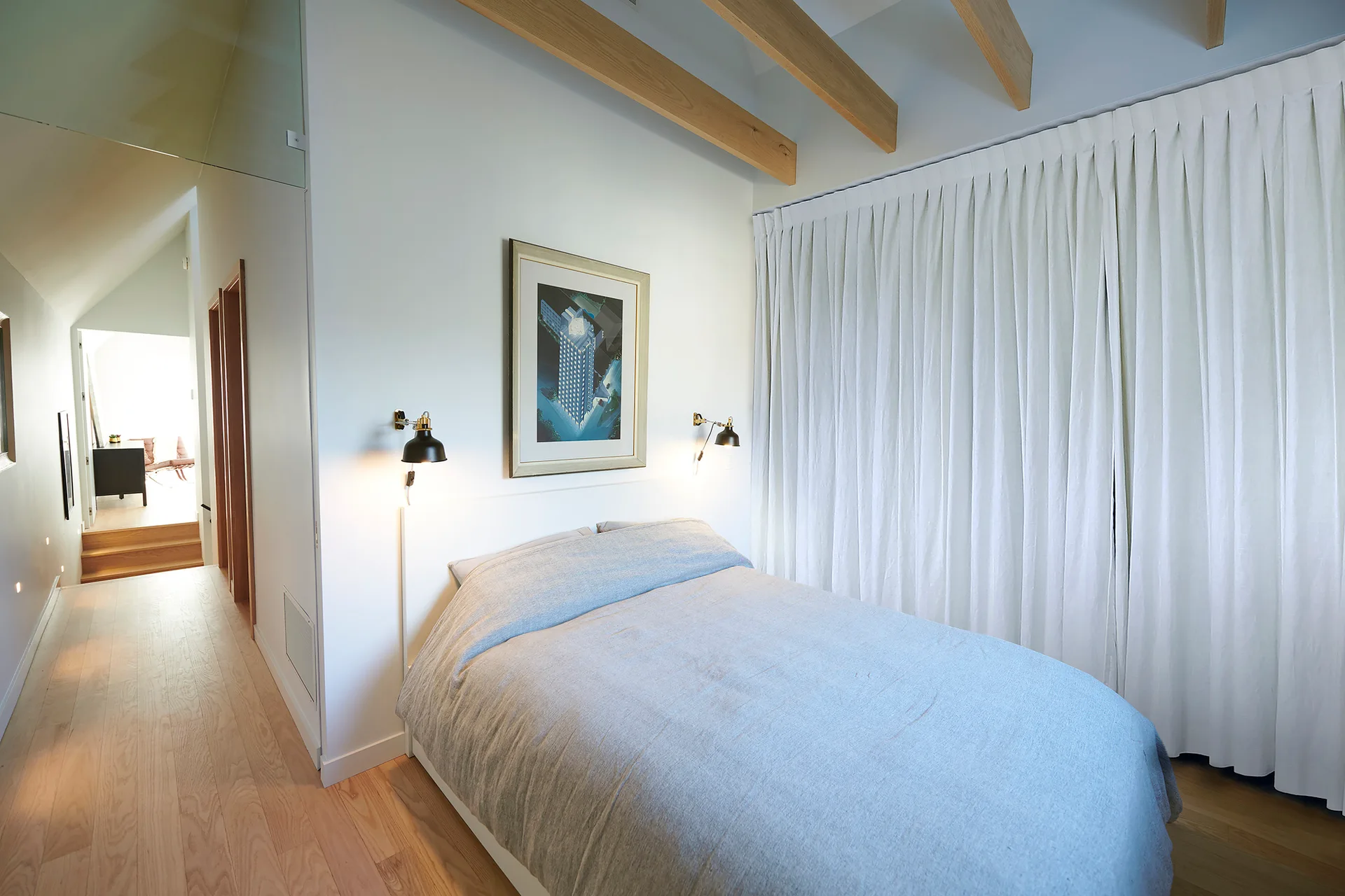 A minimalist bedroom featuring a bed with light blue linens, black wall sconces, pleated white curtains, and exposed wood ceiling beams.
