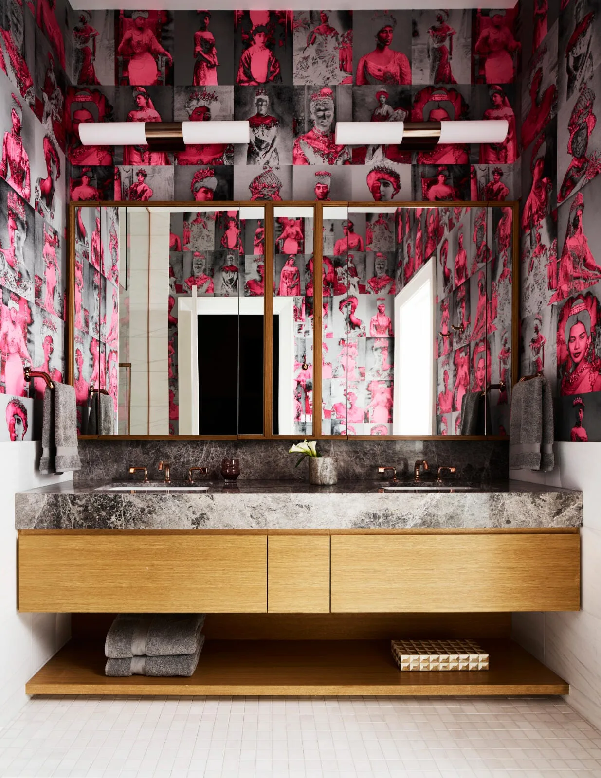 A modern bathroom features a floating wood vanity with a thick grey stone countertop beneath large mirrors and bold pink portrait collage wallpaper.