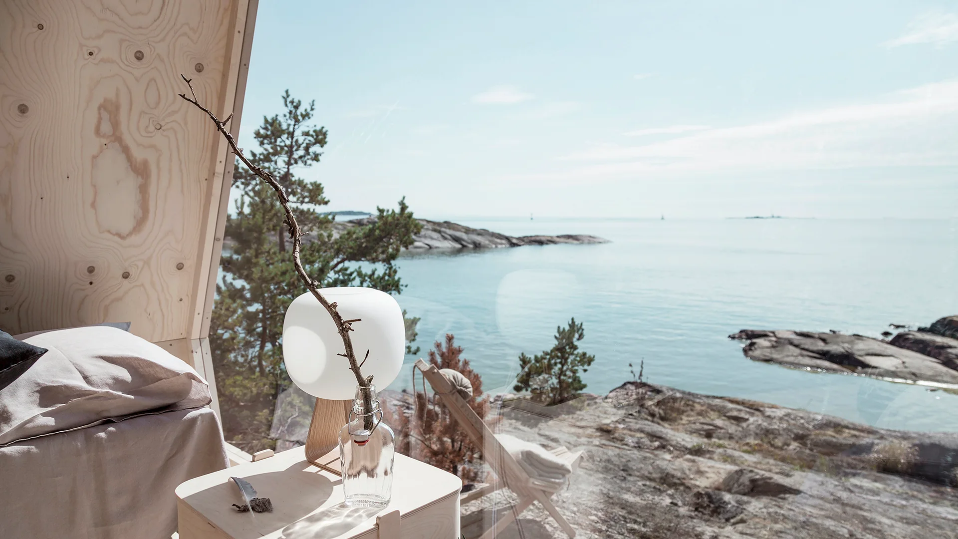 A minimalist bedroom with plywood walls, a nightstand with a globe lamp, and a large window looking out at a rocky coastline.