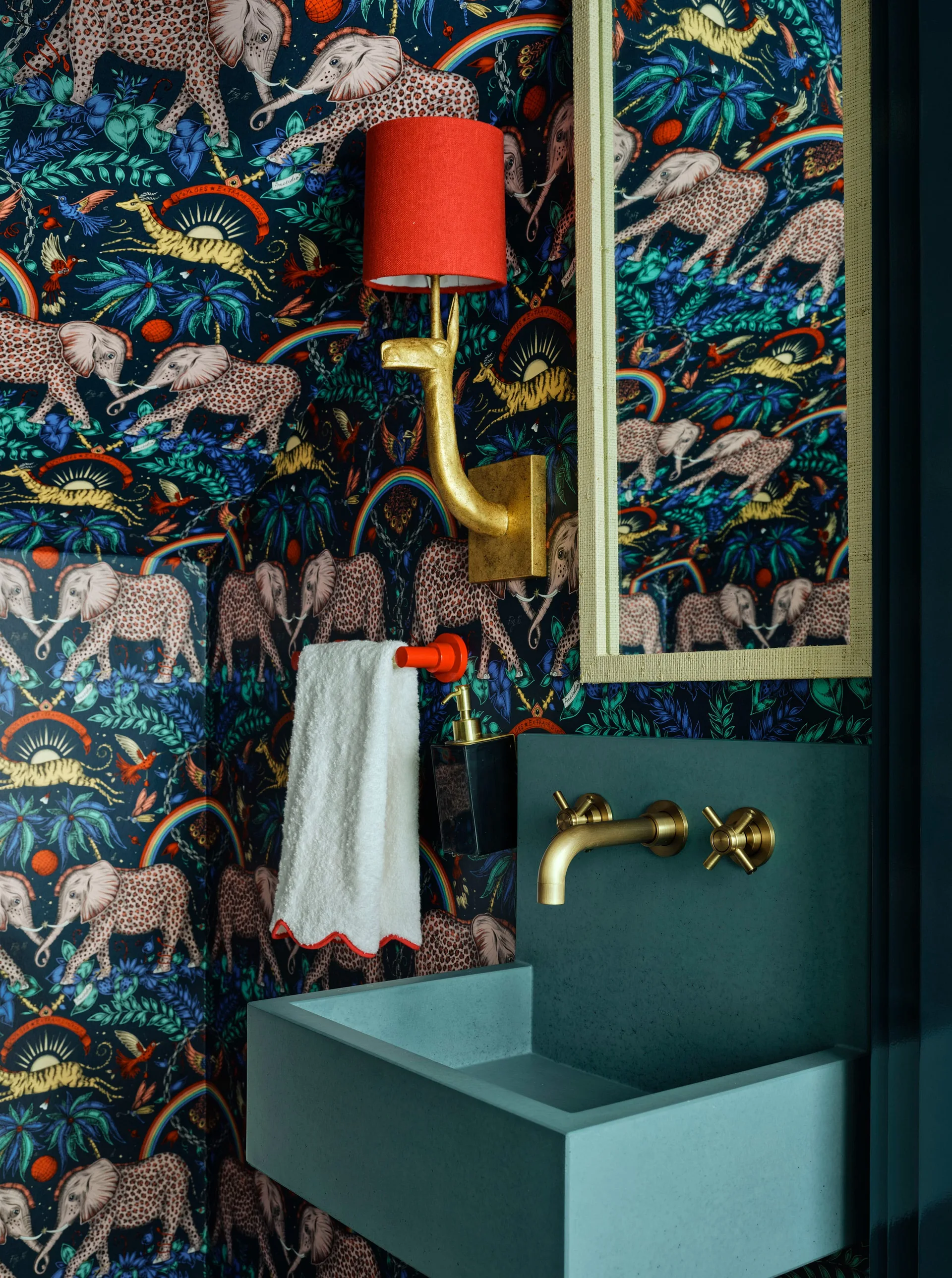 A powder room features bold elephant-patterned wallpaper, a teal wall-mounted sink with a brass faucet, and a gold animal-shaped sconce with a red shade.
