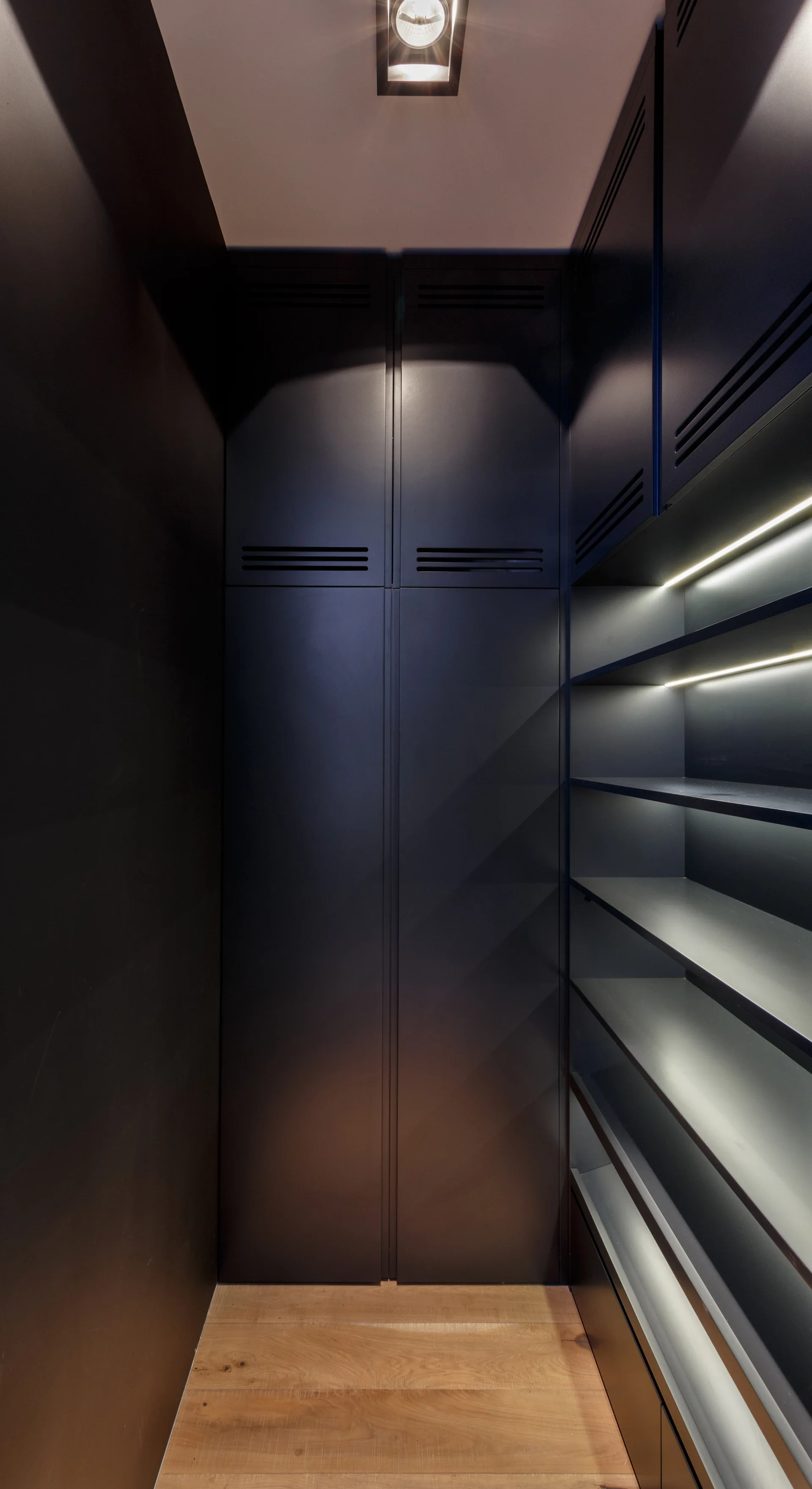 A narrow walk-in closet featuring matte black storage cabinets, open shelves lit by integrated LED strip lights, and light wood flooring.