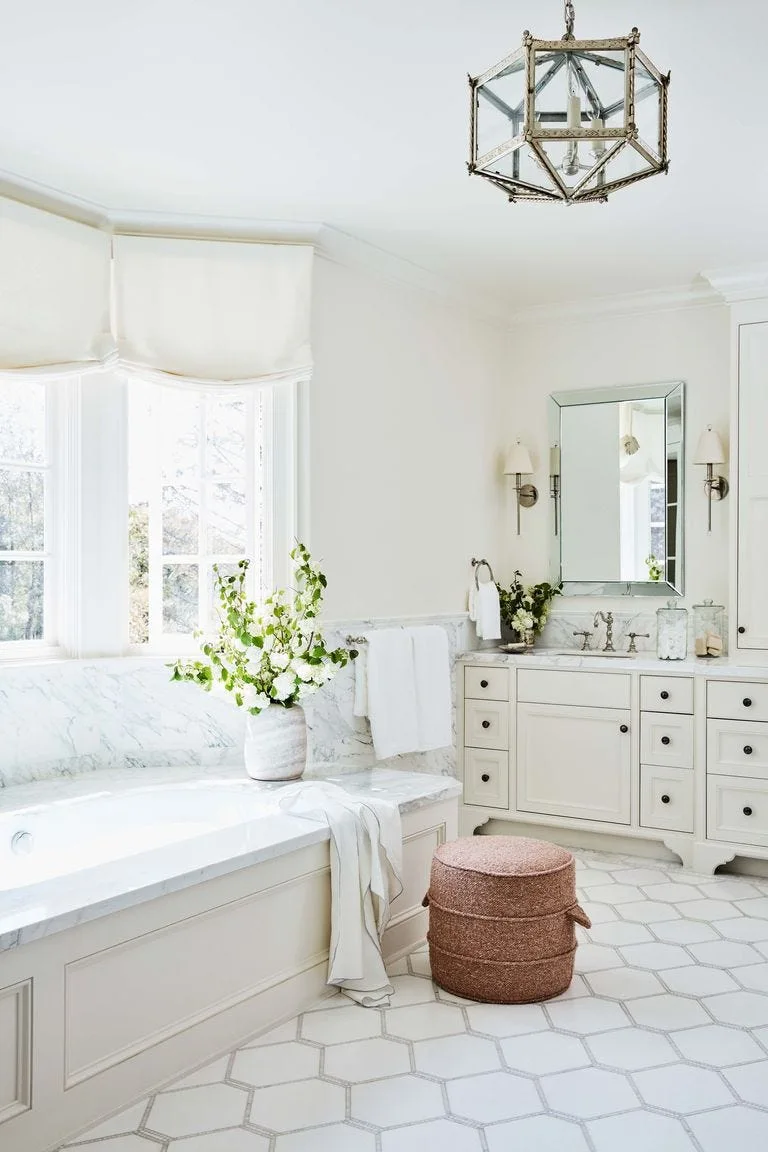 A bright bathroom features a white vanity, a marble-topped soaking tub, an earthy woven ottoman, and white hexagonal floor tiles.