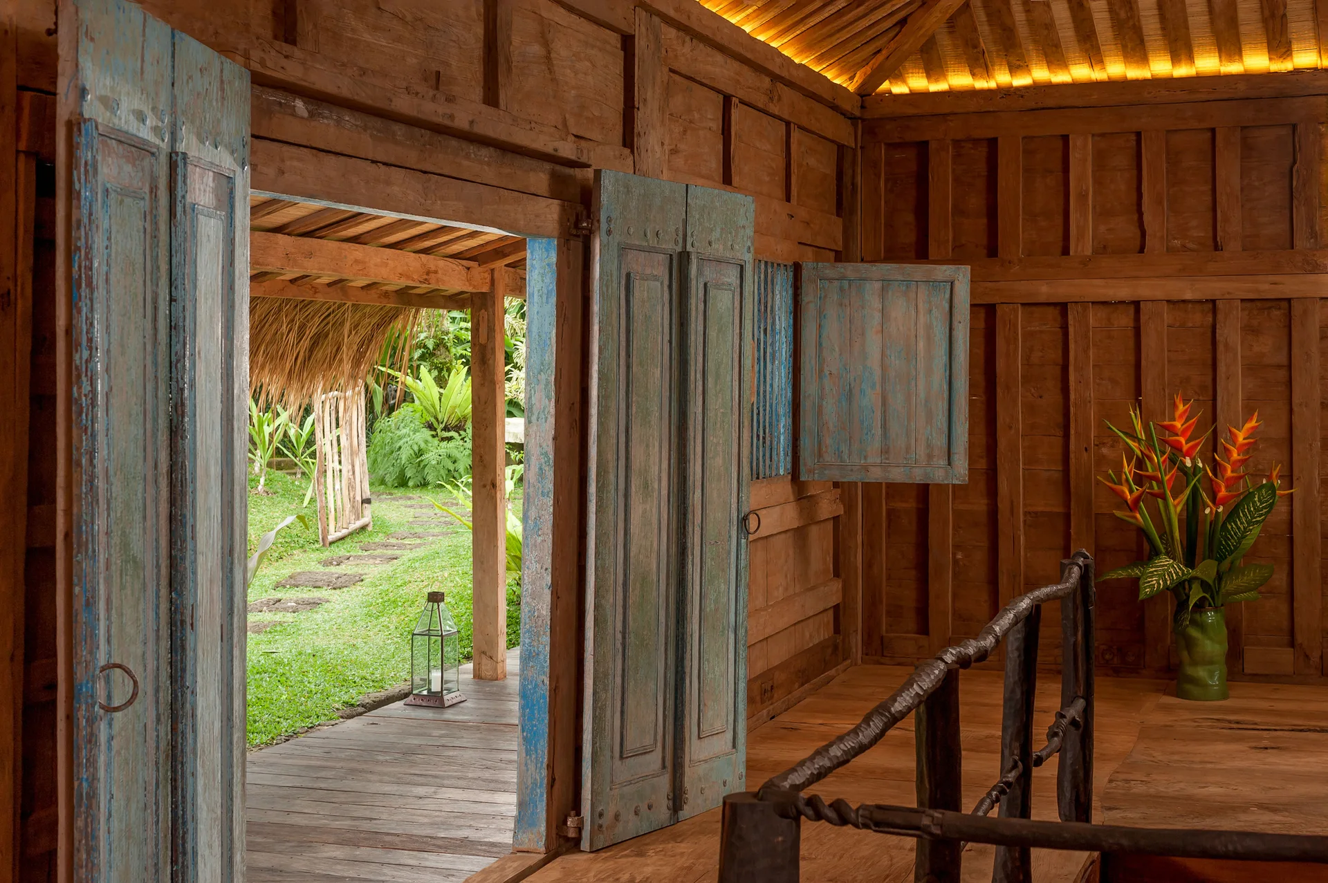 A rustic wood-paneled interior features distressed blue doors opening to a lush green garden and a dark twisted wood railing.