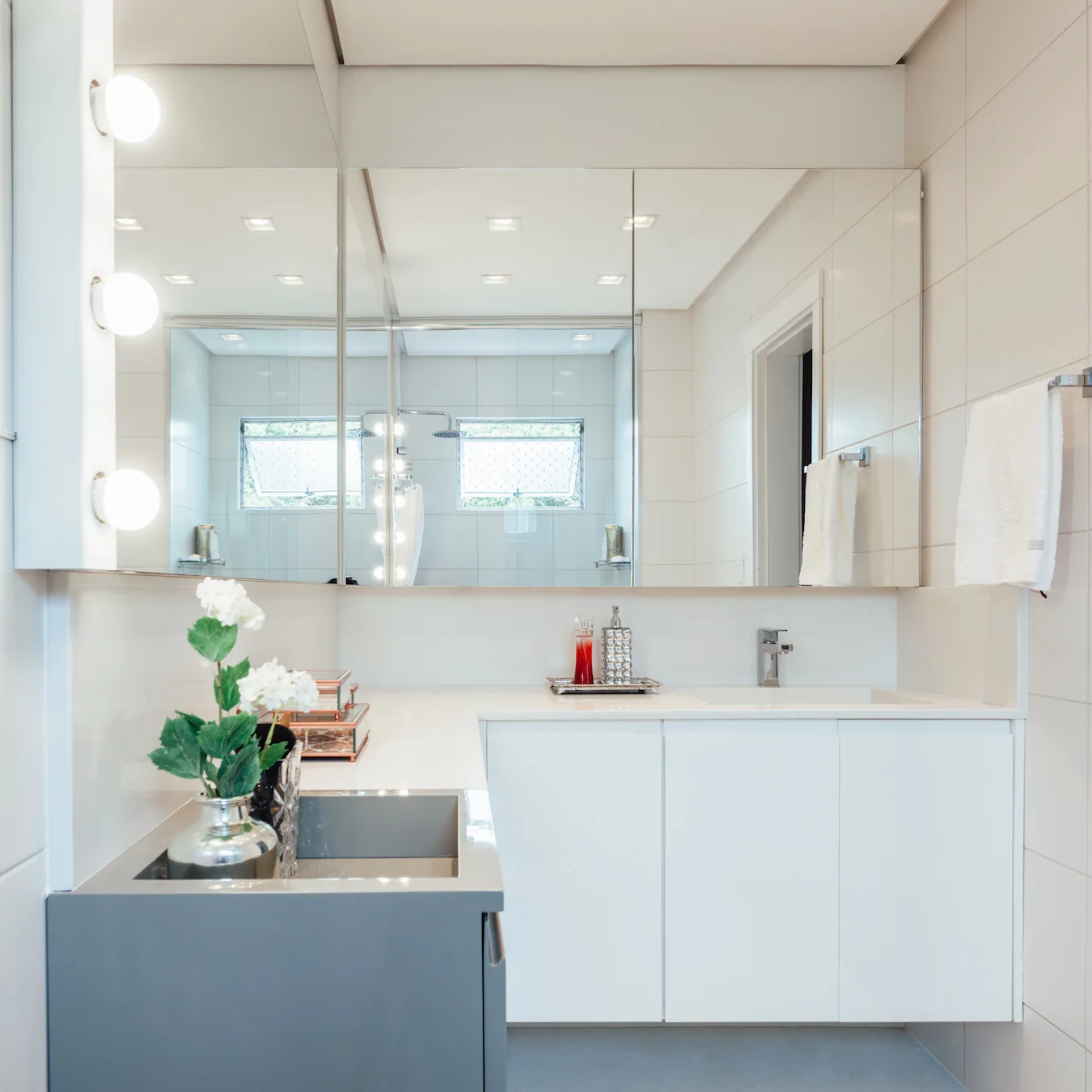 A contemporary bathroom vanity features white and grey floating cabinets, a large frameless mirror with bulb lights, and a white tiled wall.