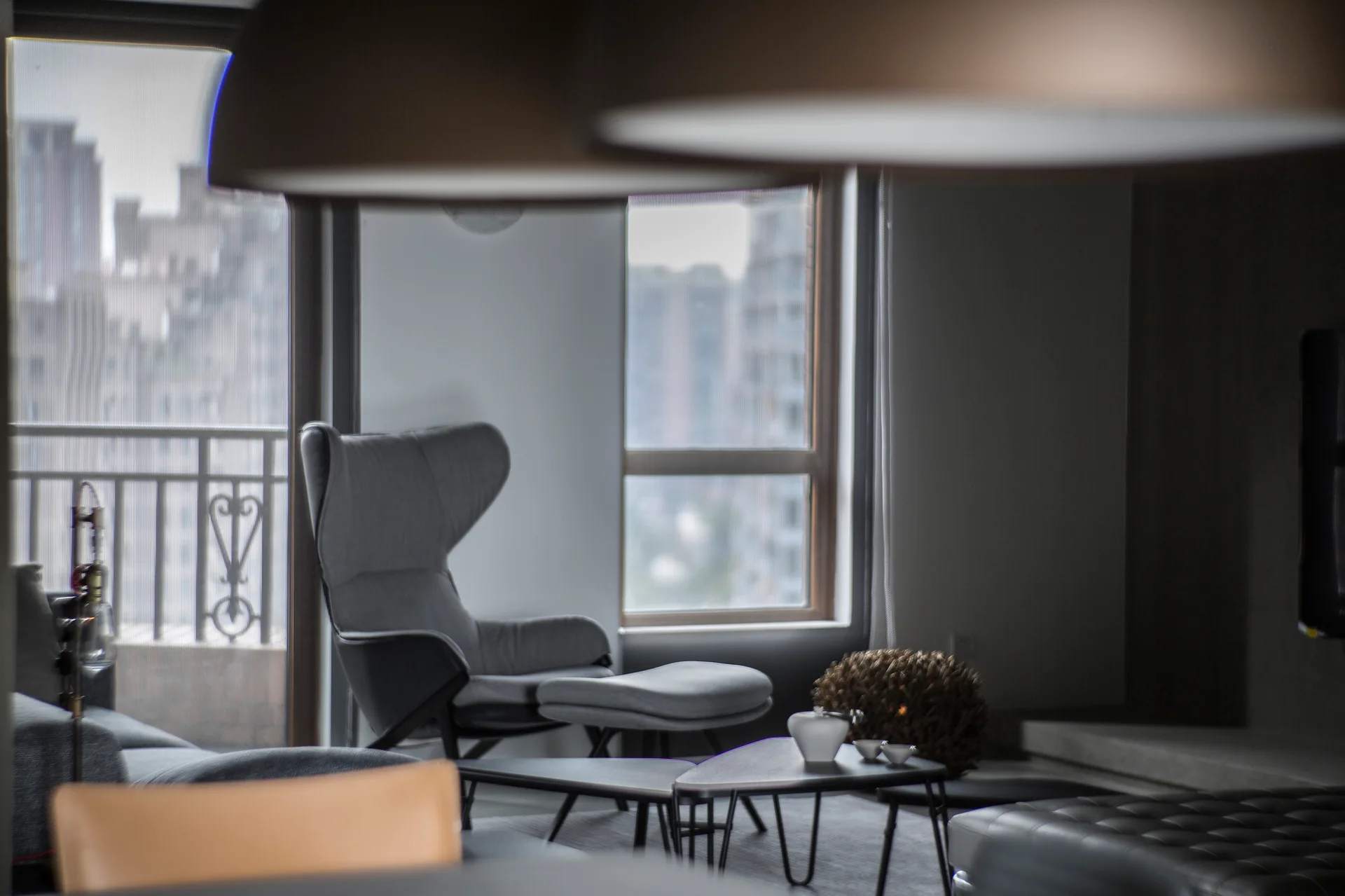 A grey wingback lounge chair and matching ottoman sit near a large window overlooking a city, with low-profile coffee tables in the foreground.