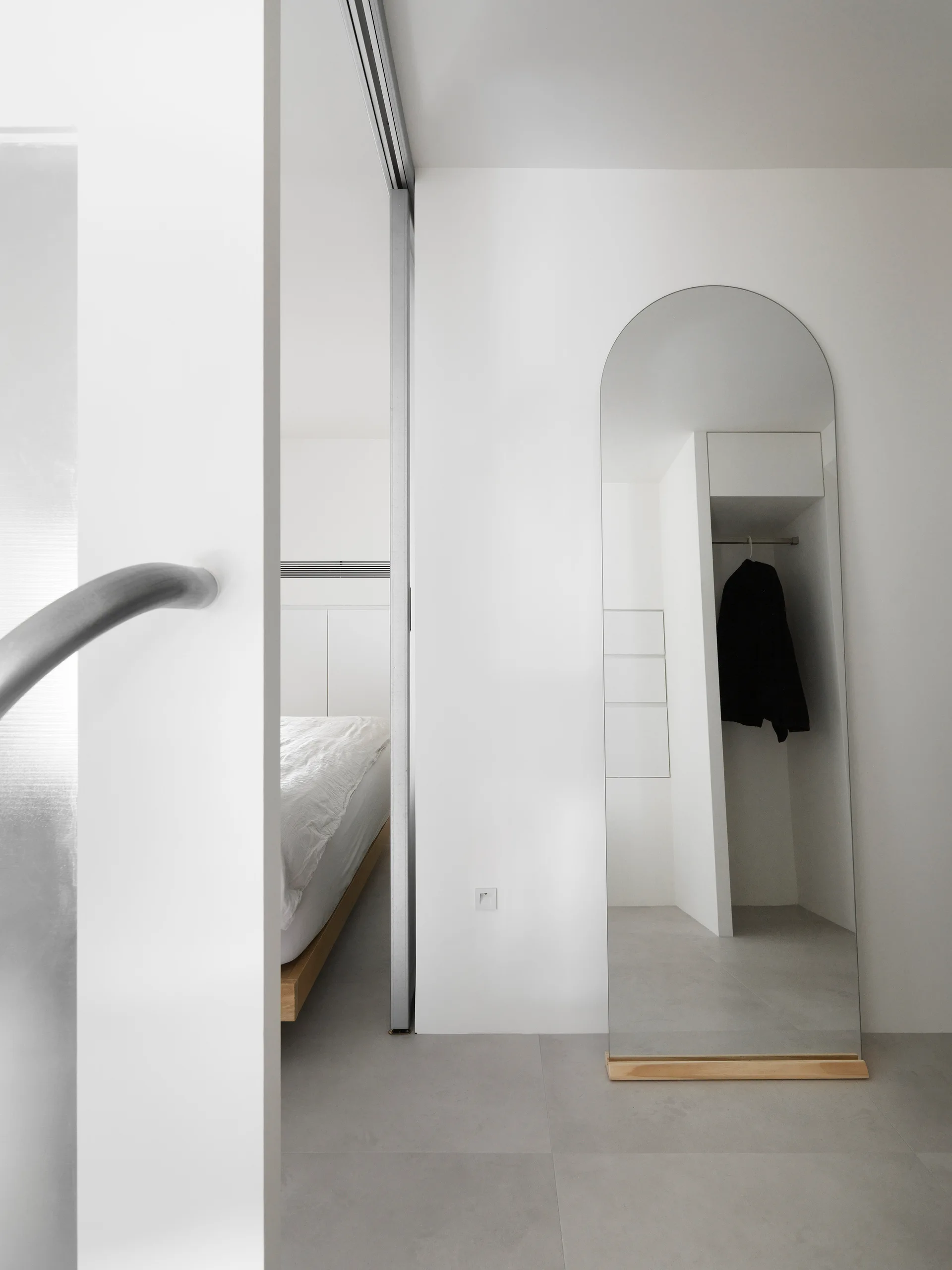 A minimalist white bedroom entryway features a sliding door, light grey tile flooring, and a tall arched floor mirror in a light wood base.