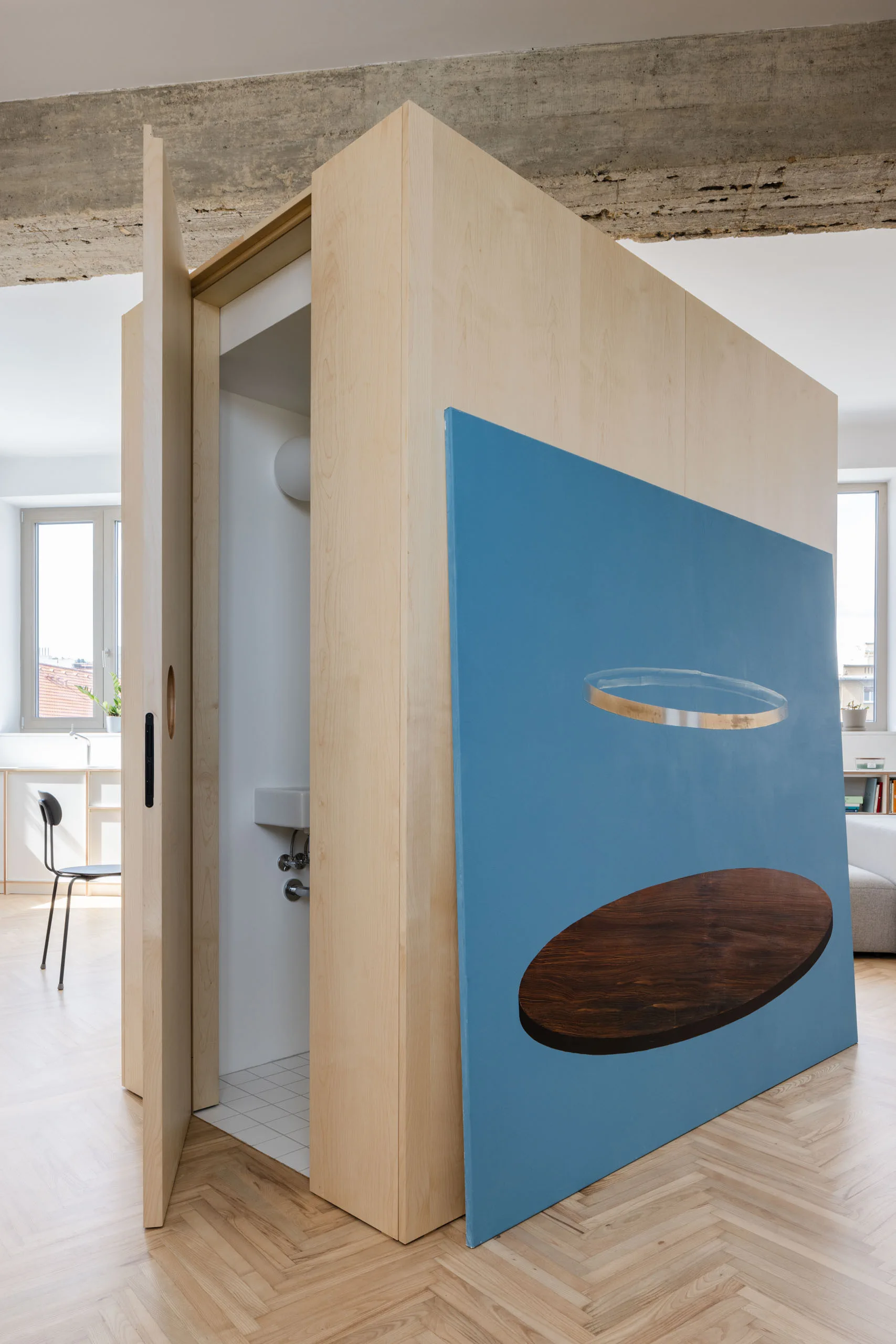 A light plywood partition containing a tiled bathroom stands on herringbone flooring next to a large blue painting.