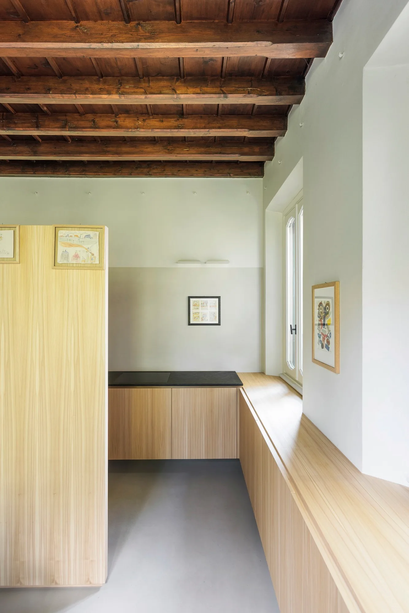 A minimalist kitchen features light wood cabinets beneath a window, a dark countertop, grey flooring, and dark rustic wood ceiling beams.