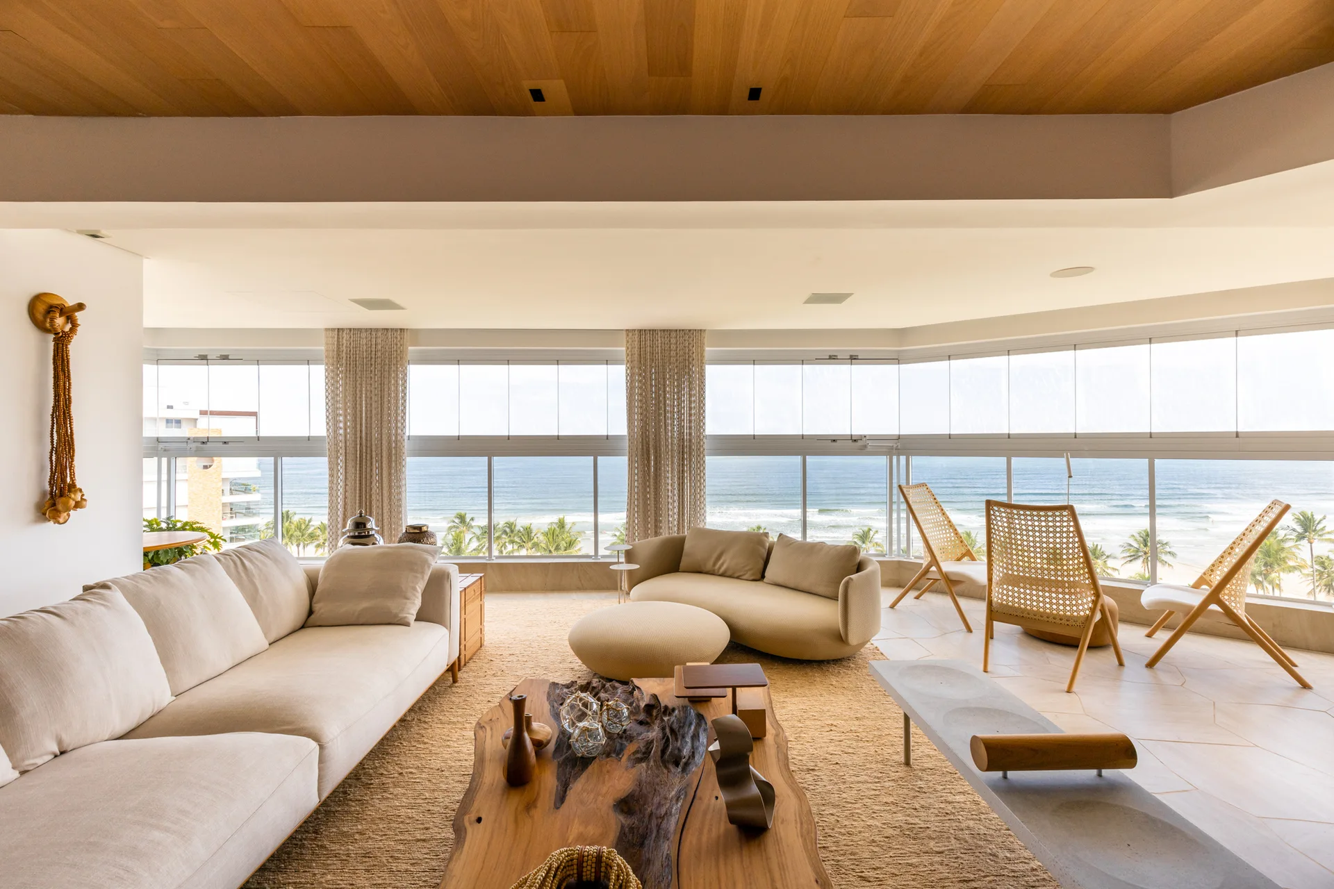A coastal living room features a cream sofa, an organic wood slab coffee table, and rattan chairs facing large windows with an ocean view.