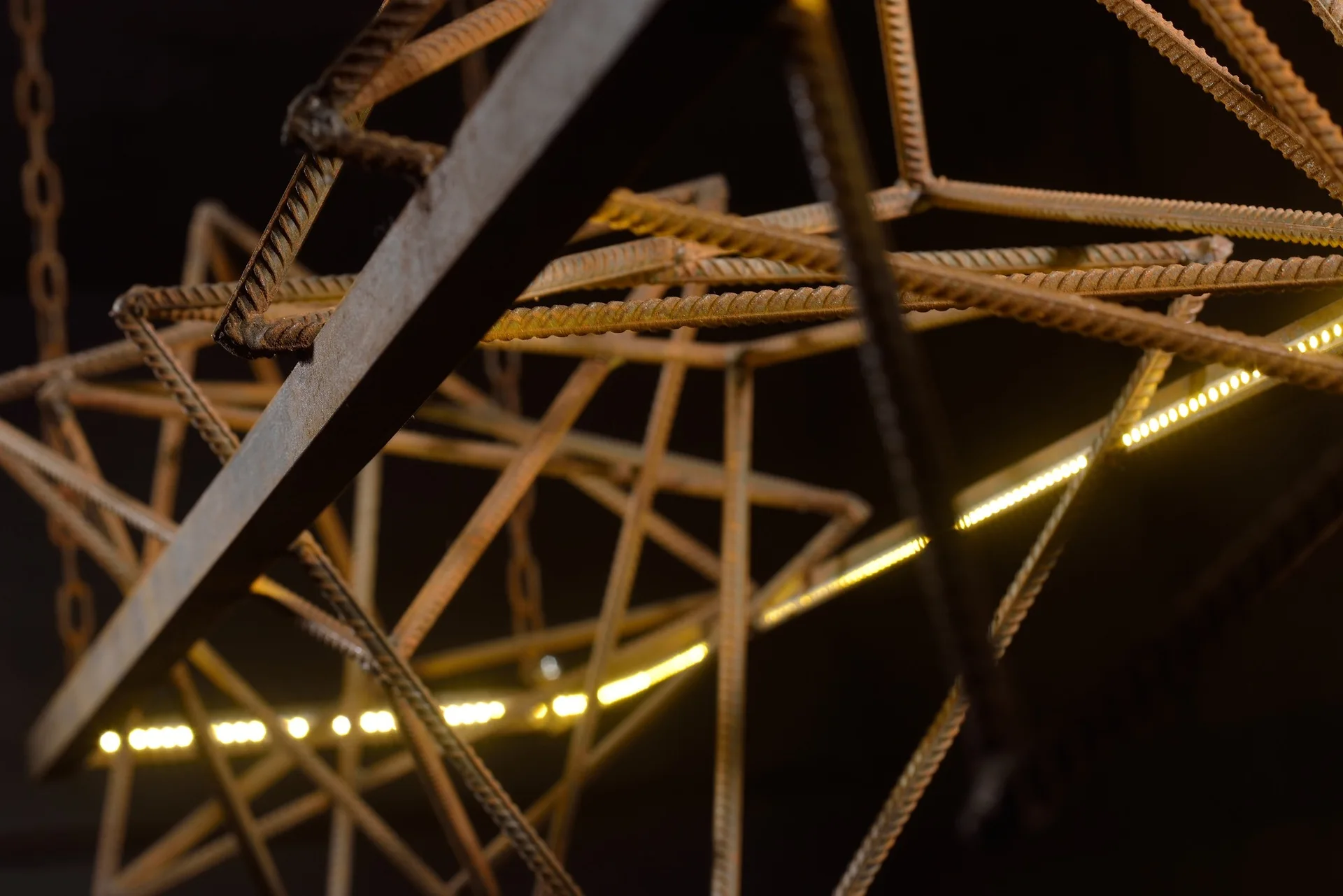 An industrial geometric pendant light crafted from rusted metal rebar features integrated warm yellow LED strip lighting against a dark backdrop.