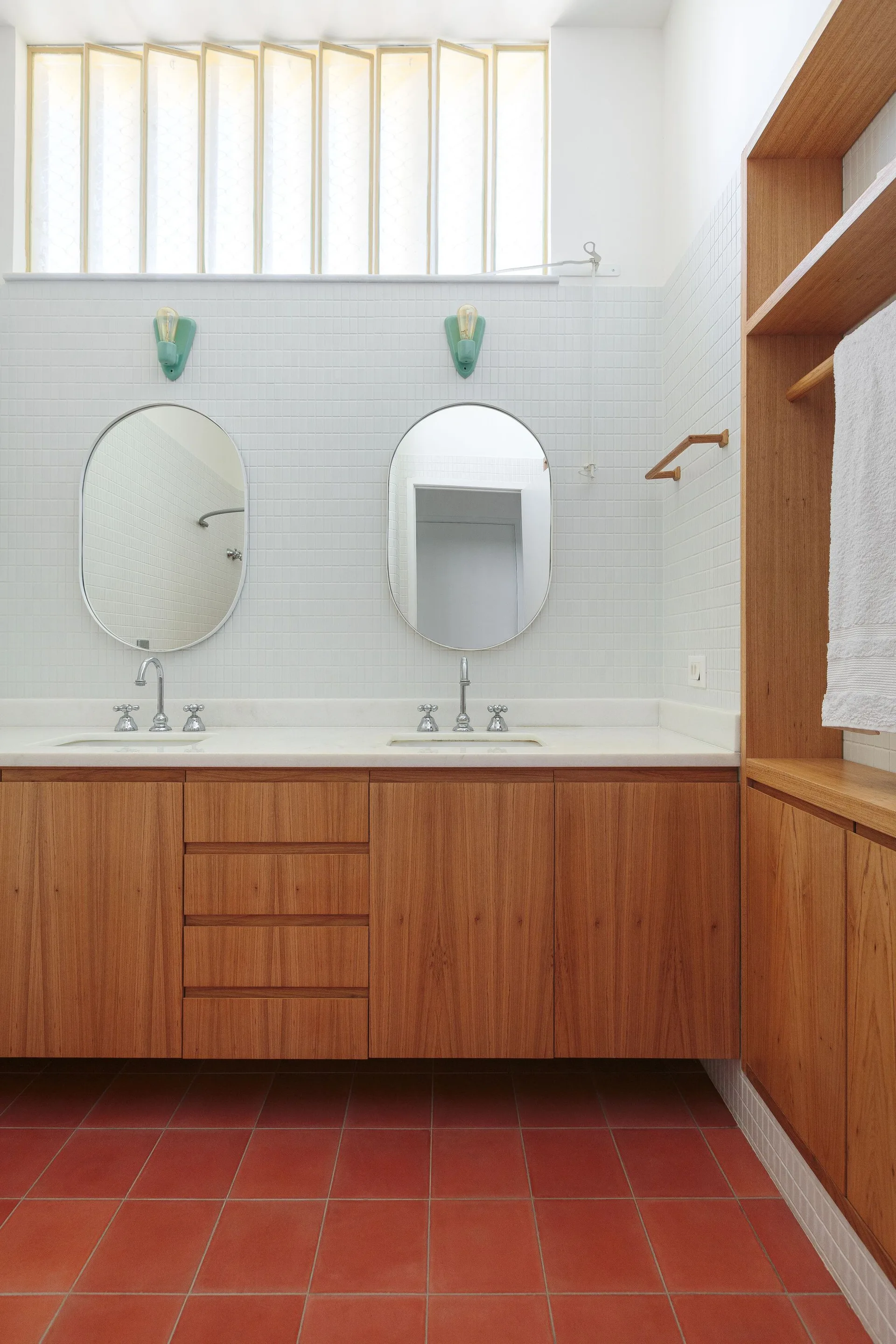 A bright bathroom features a wooden double vanity with oval mirrors against white grid tiled walls and red terracotta floor tiles.