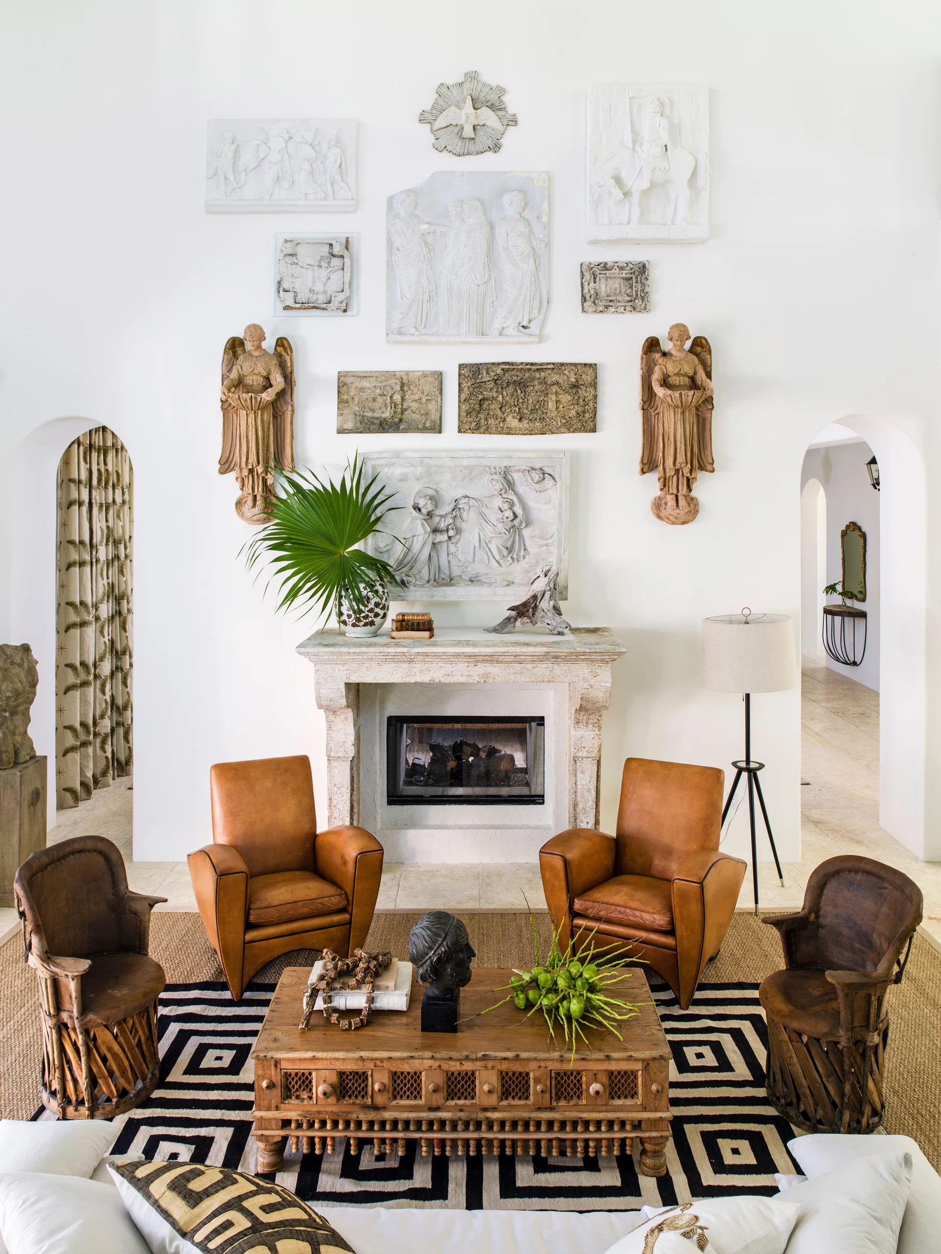A living room features a stone fireplace, tan leather armchairs, and a symmetrical gallery wall of white plaster reliefs under a high ceiling.