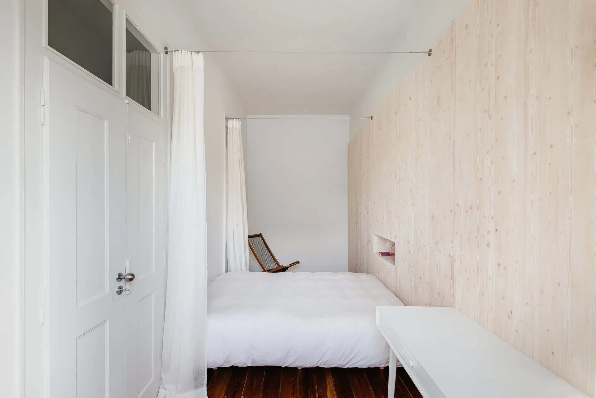 A narrow bedroom features a bed with white linens set against a light wood paneled wall with a small recessed niche.