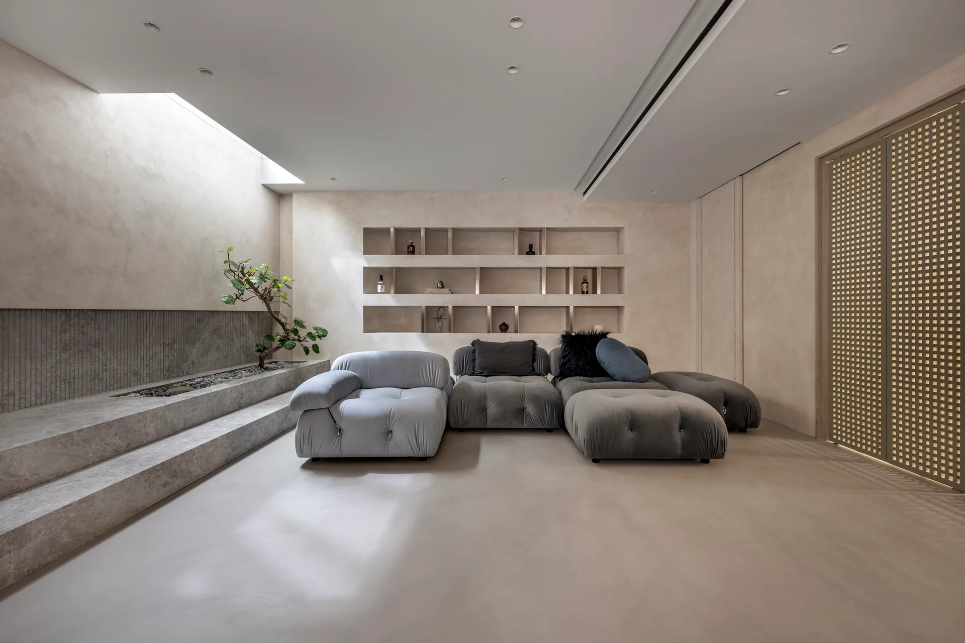 A minimalist living room features a low grey modular sofa, built-in wall shelving, and a tiered stone platform planter under a skylight.