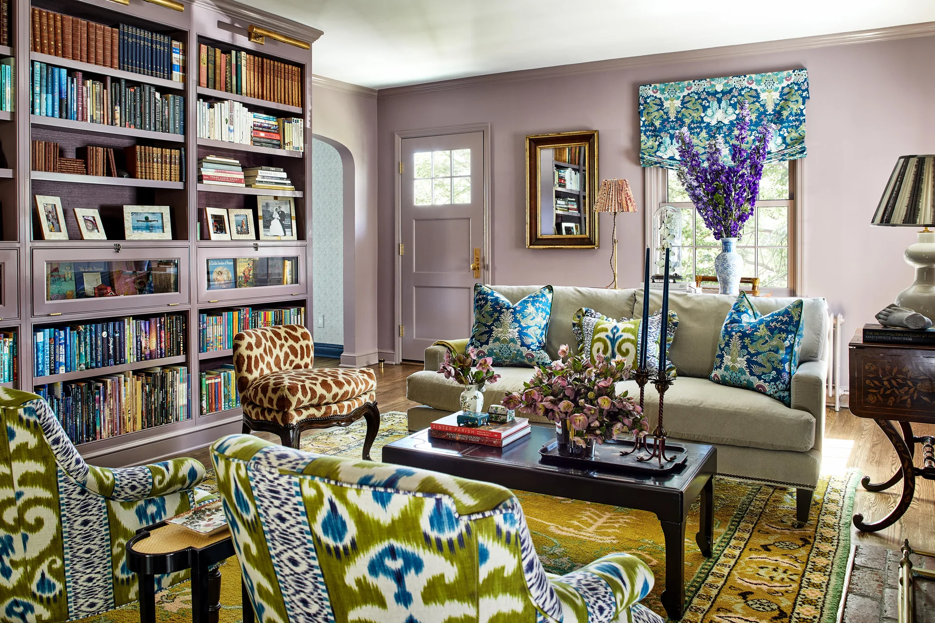 A traditional living room featuring a lavender built-in bookcase, lavender walls, a light beige sofa, and blue-and-green patterned armchairs.