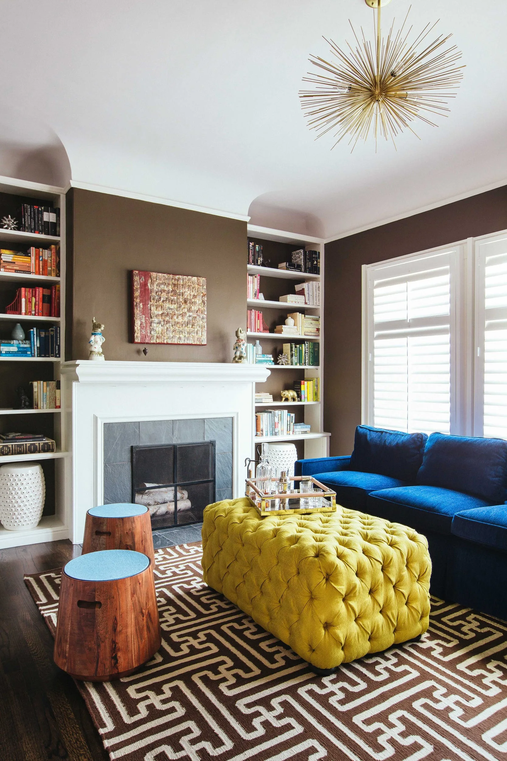 A living room featuring chocolate brown walls, a blue sofa, a tufted yellow ottoman, and white bookshelves flanking a fireplace.