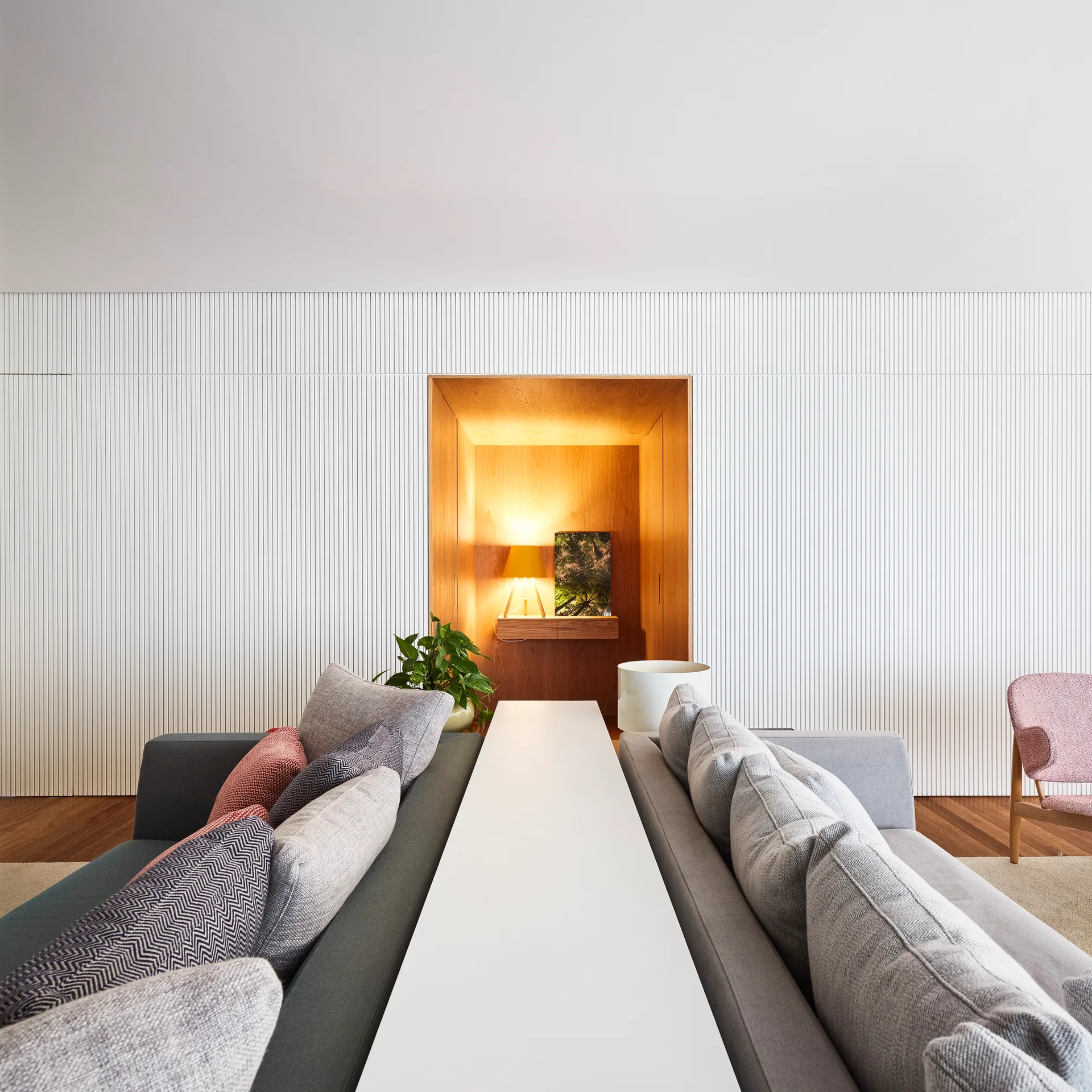 Two gray sofas sit back to back separated by a long white console table against a white slatted wall with a recessed warm wood alcove.