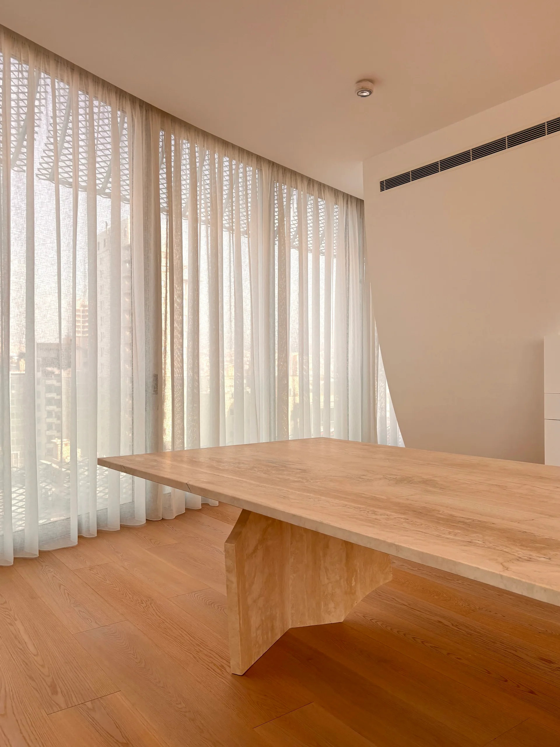 A large stone dining table sits on light wood flooring in a minimalist room with floor-to-ceiling sheer cream curtains.
