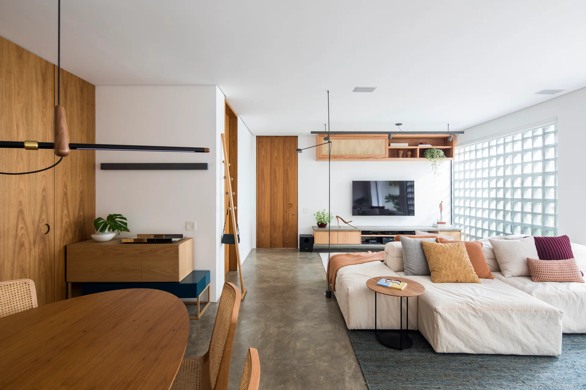 A modern open-plan living room featuring a cream sectional sofa, a glass block window wall, polished concrete floors, and warm wood wall paneling.