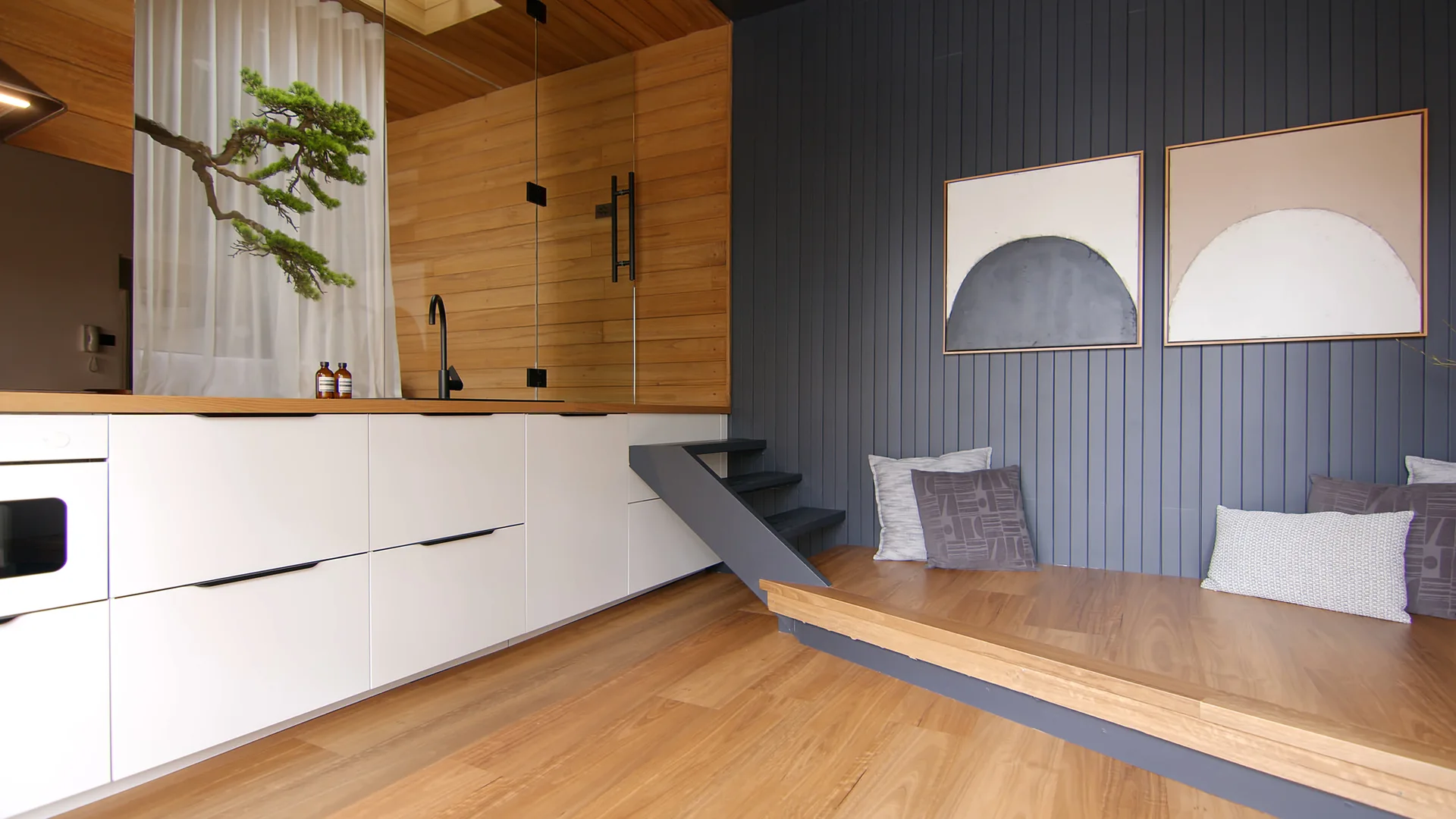A modern kitchen with white cabinets and a black faucet next to a raised wooden seating platform with grey wood paneled walls.