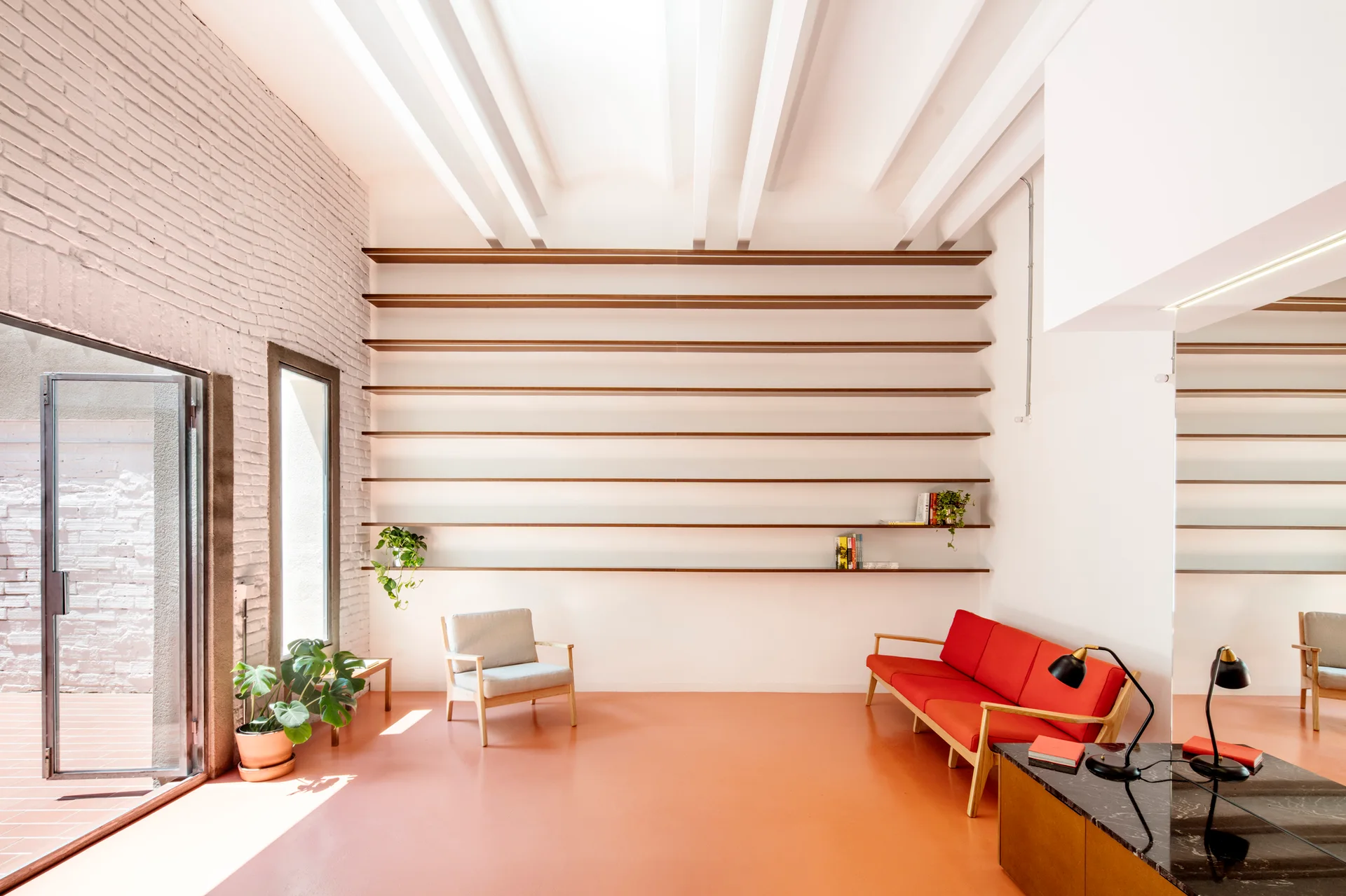 A bright living space features a red mid-century sofa, a grey armchair, and long horizontal wood shelves on a white brick wall above a terracotta floor.