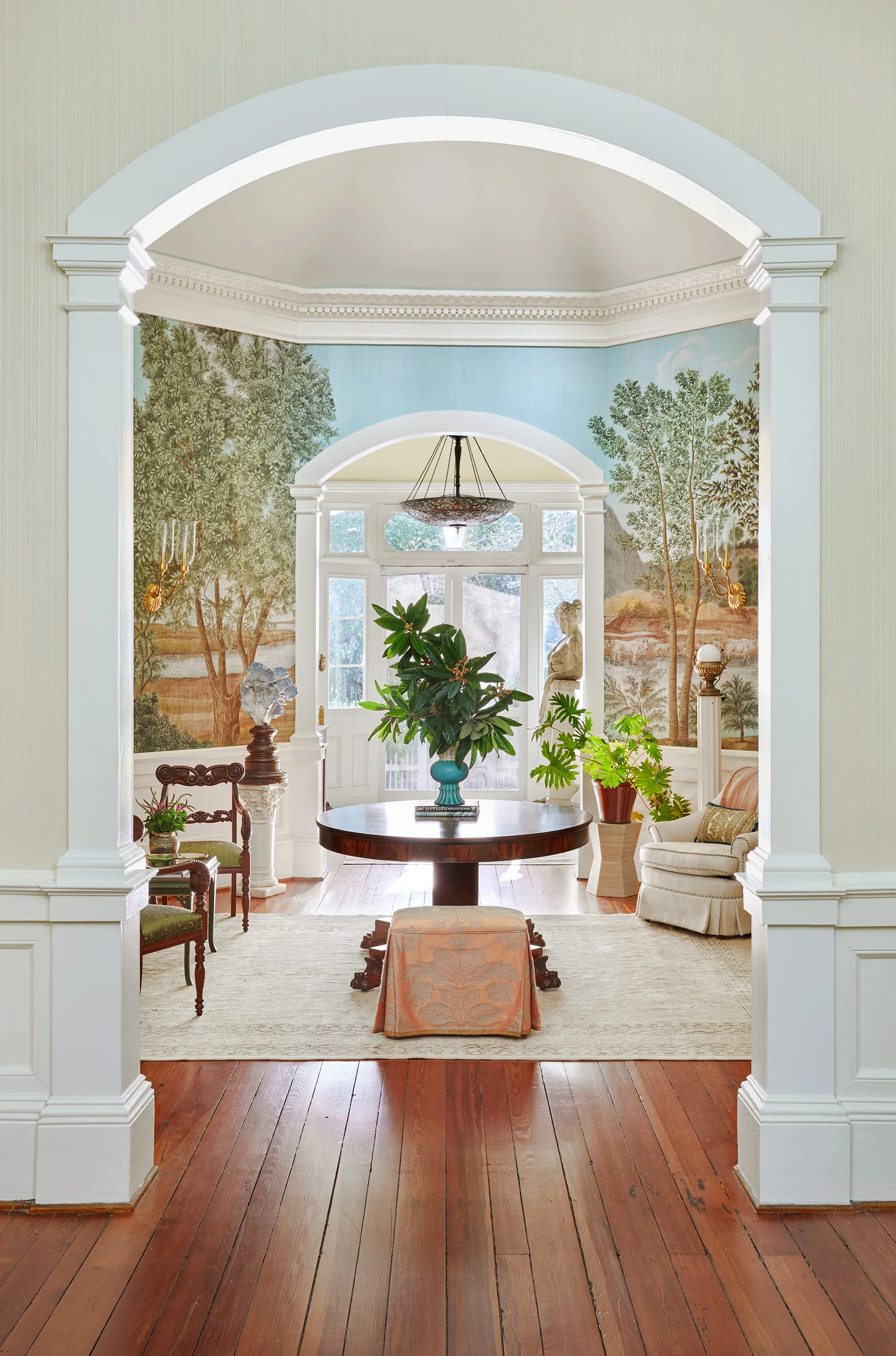 A formal foyer viewed through a white archway featuring a central round wooden table with green foliage, scenic landscape mural wallpaper, and hardwood floors.