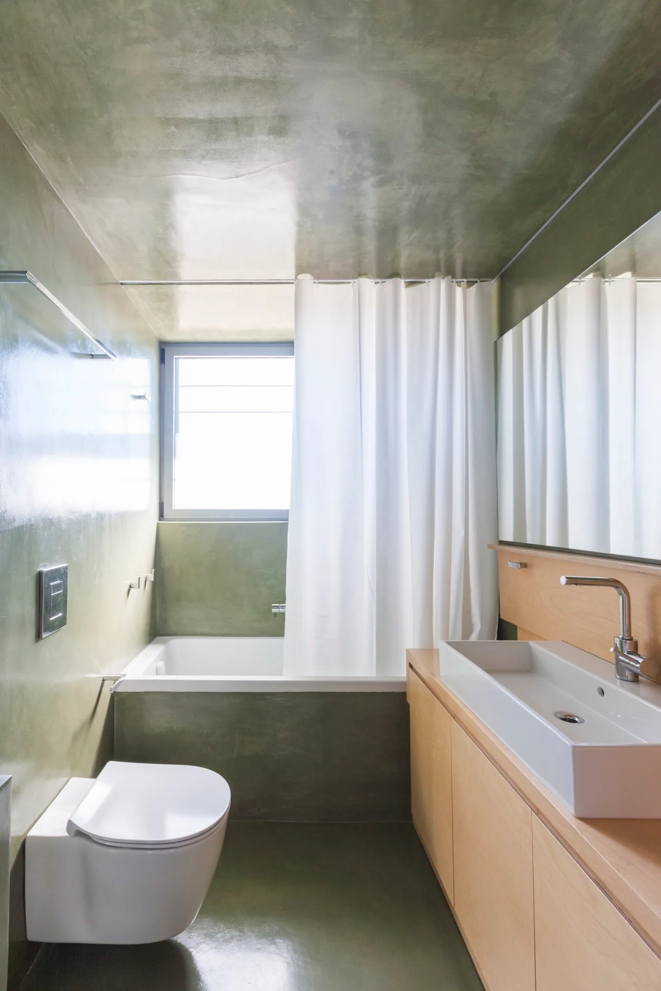 A modern minimalist bathroom featuring glossy olive green plaster walls, a floating white toilet, and a light wood vanity with a white ceramic sink.