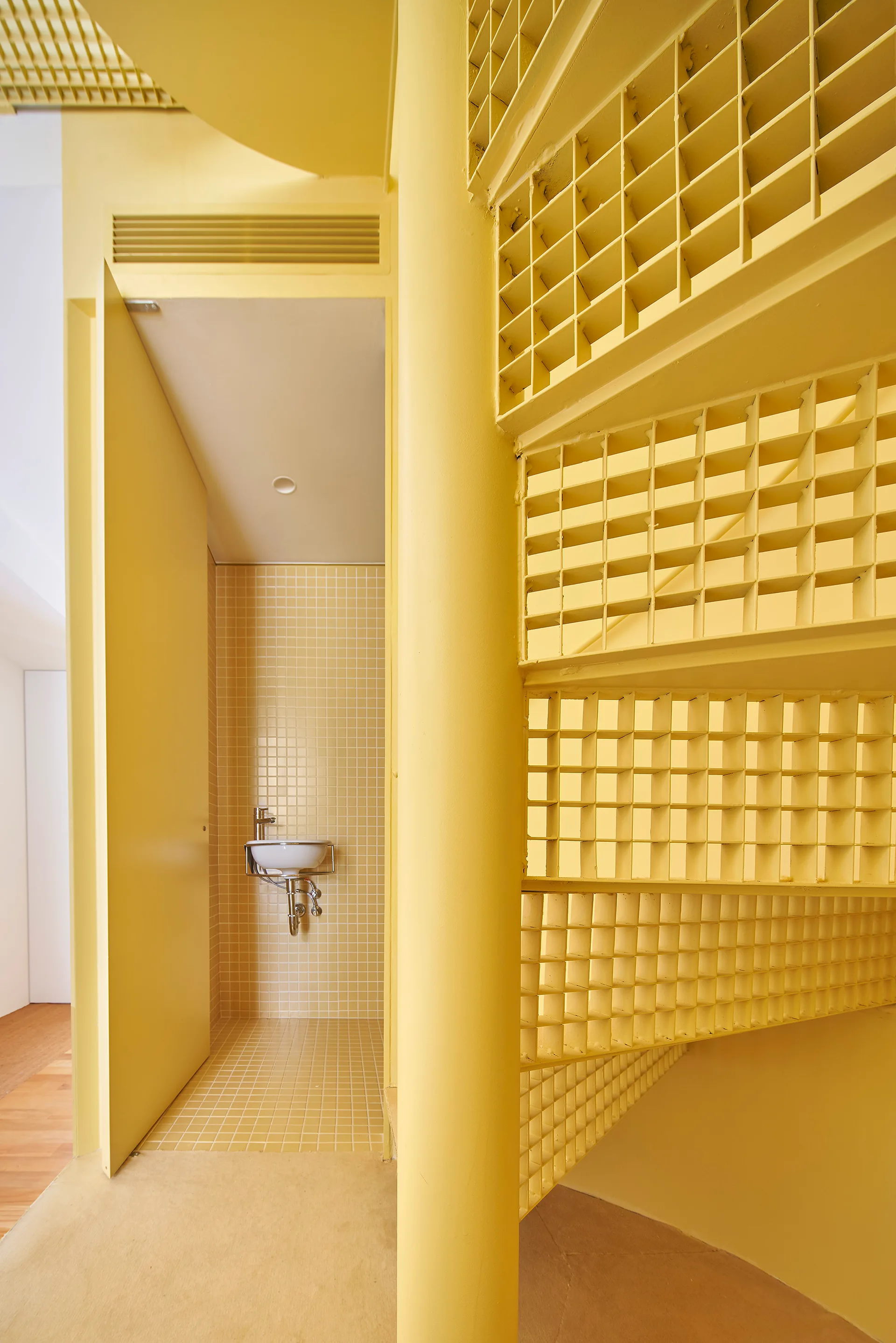 A bright yellow metal spiral staircase with grid railings stands next to a small, tiled alcove containing a white wall-mounted sink.