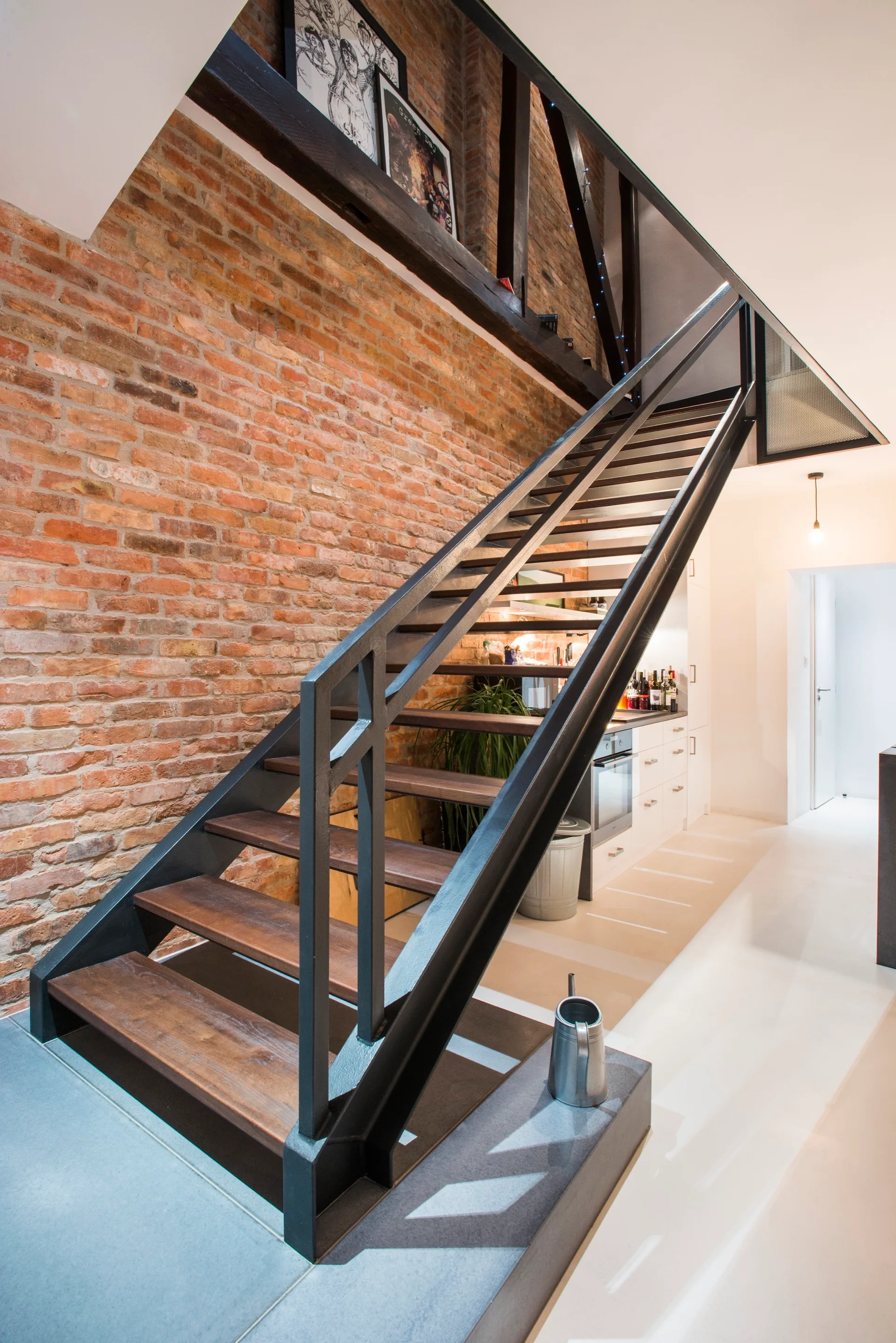 An open-riser industrial staircase with dark metal framing and wooden treads rises against a rustic terracotta exposed brick wall.