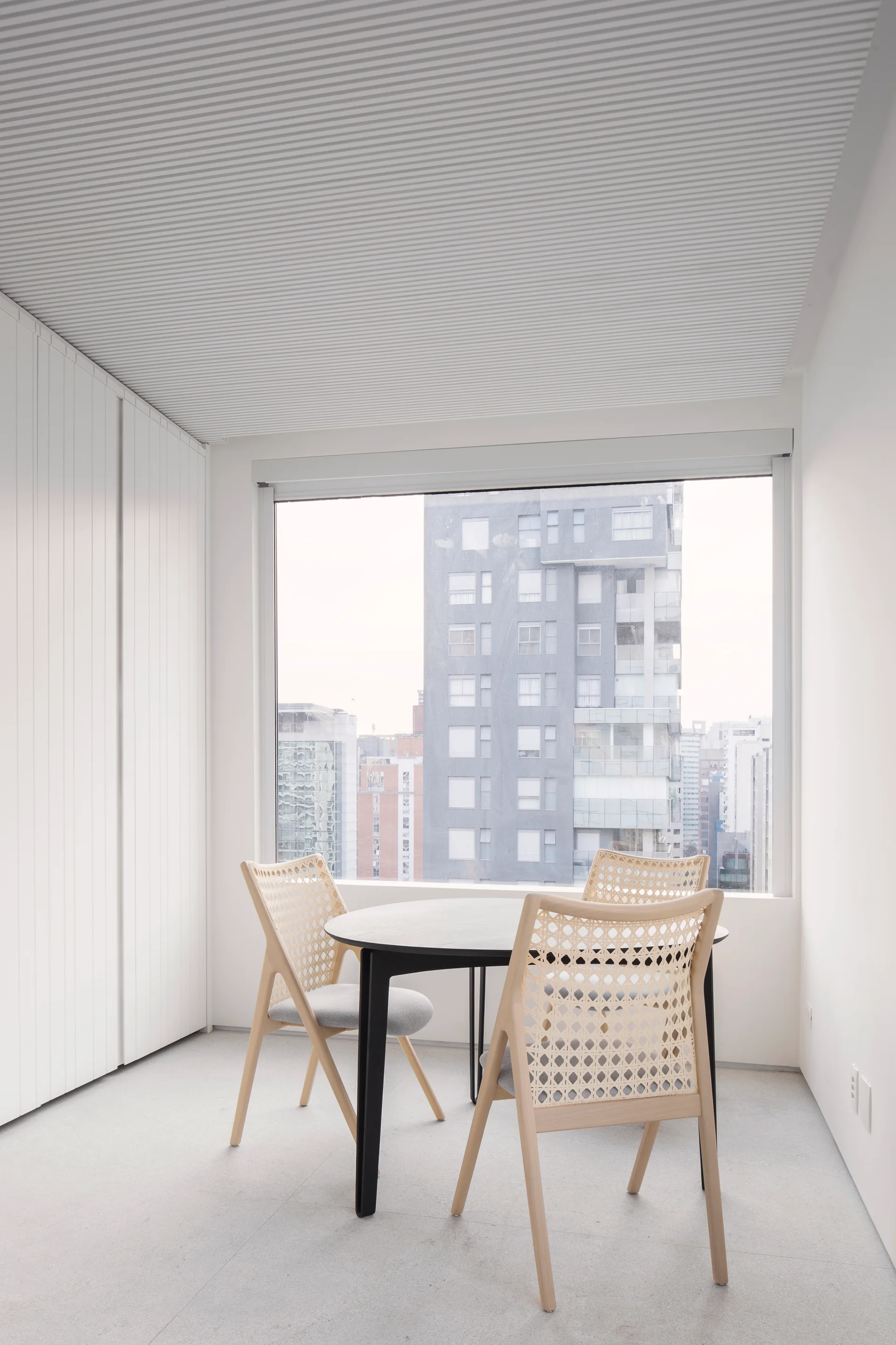 A round black dining table and light wood cane back chairs sit in a minimalist white room with a ribbed ceiling and a large window.