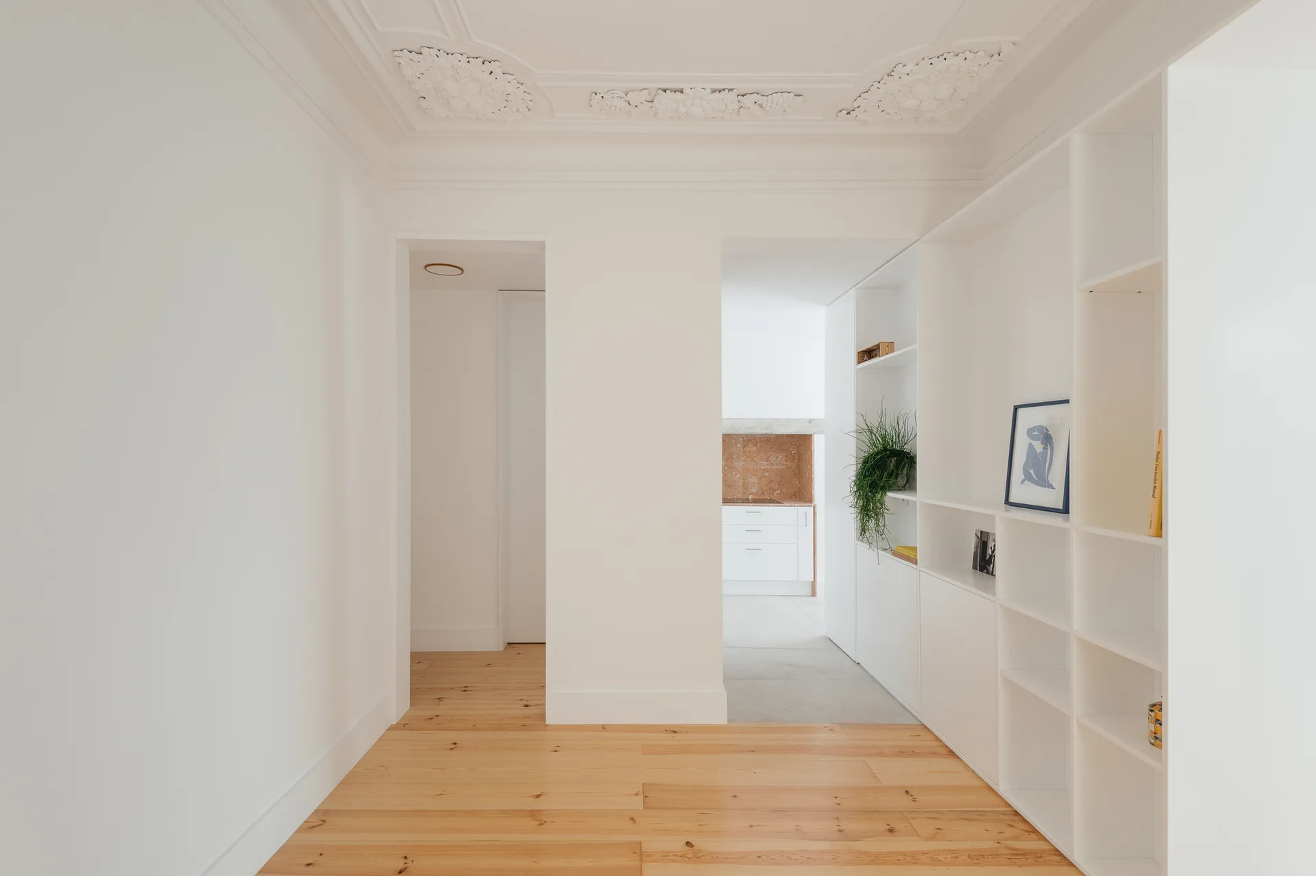 A bright white hallway features natural light wood floors, a built-in modular shelving unit, and a highly detailed ornate plaster ceiling.