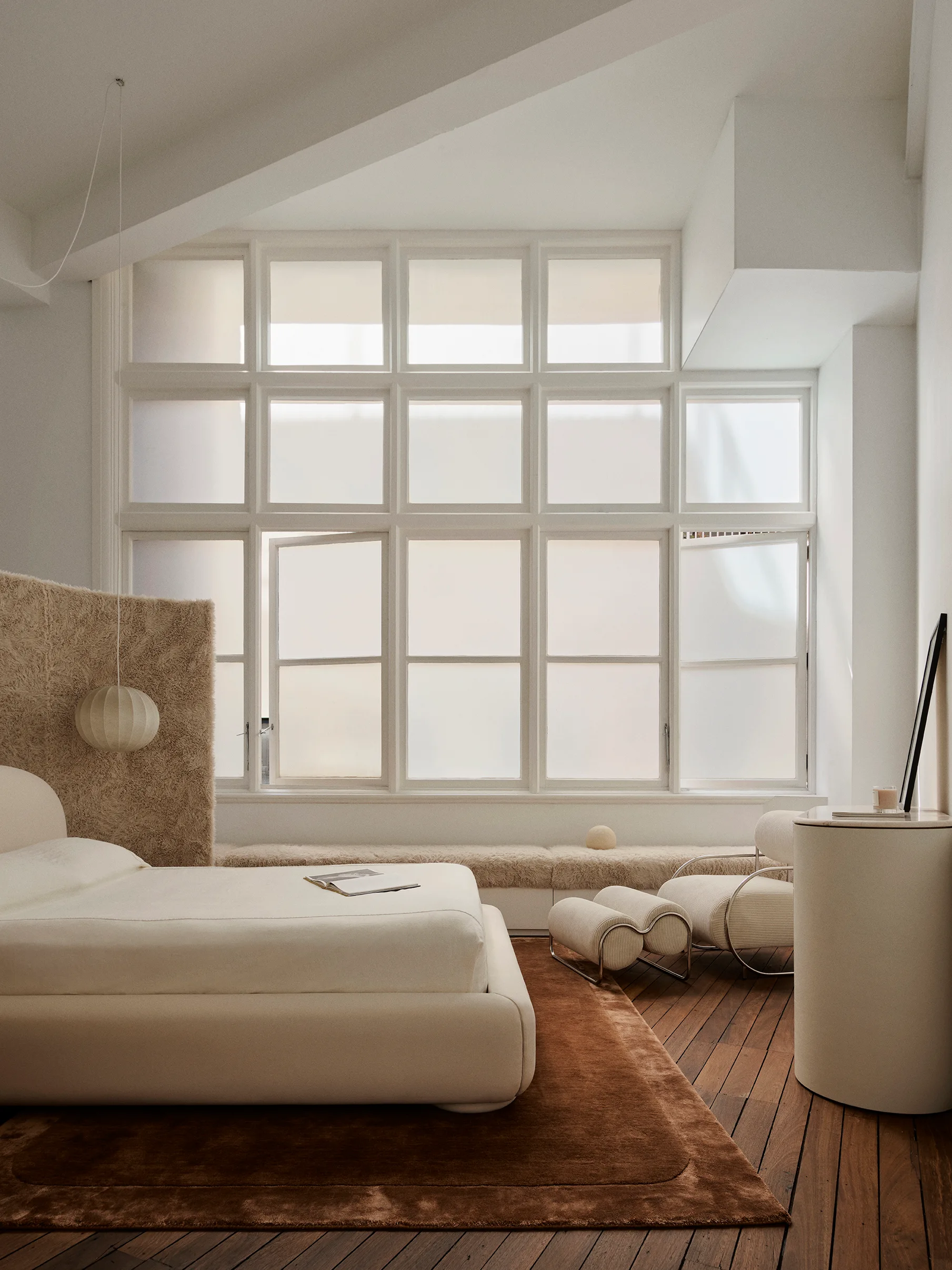 A minimalist bedroom features a low-profile cream bed, a high-pile shag room divider, a curved armchair, and a large frosted grid window.