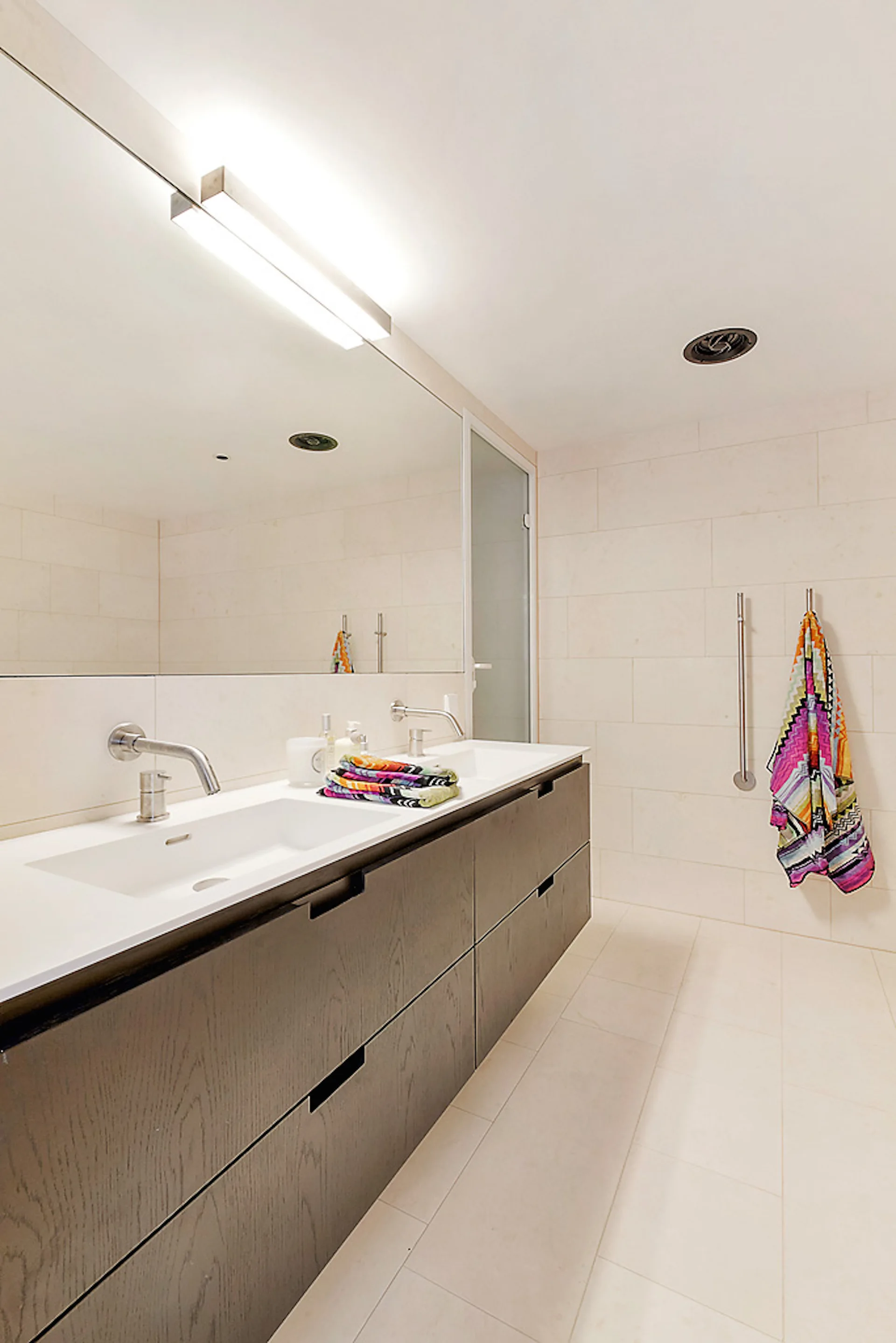 A modern bathroom featuring a dark wood floating vanity with two integrated sinks, wall-mounted faucets, a large mirror, and off-white tiled walls.