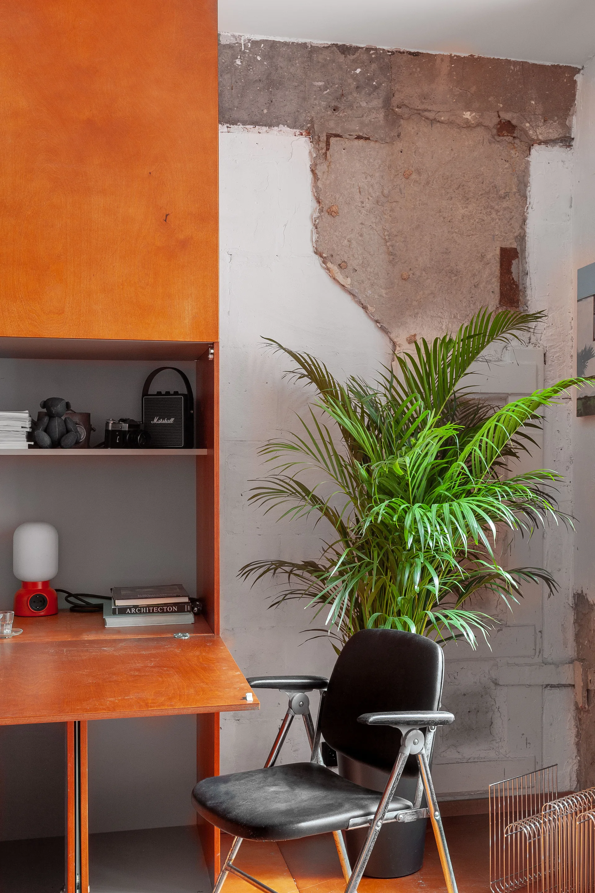 An orange plywood desk unit and a black office chair stand against an unfinished white plaster wall with a large patch of exposed masonry.