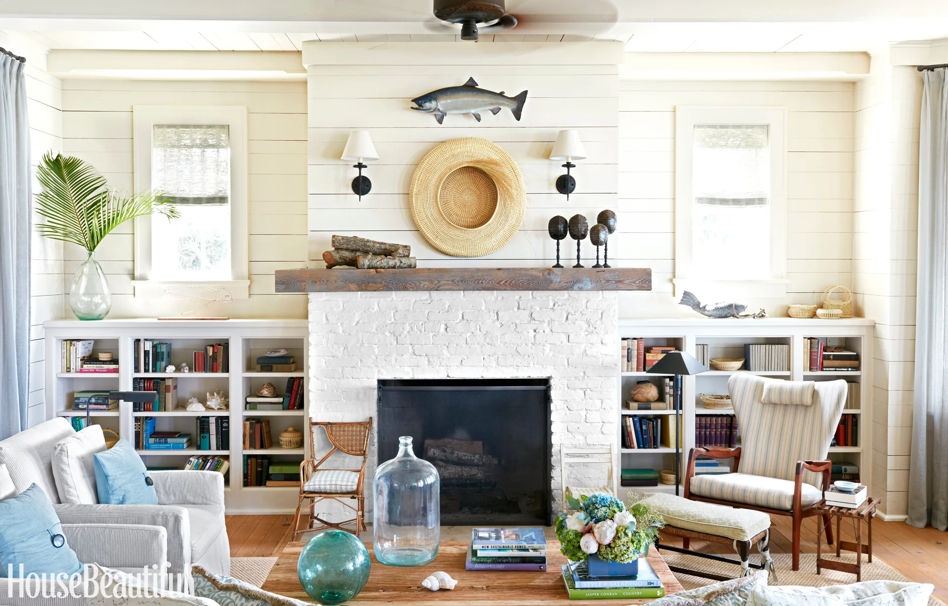 A bright living room featuring a white brick fireplace flanked by built-in bookcases, shiplap walls, and coastal decor accents like a woven hat.
