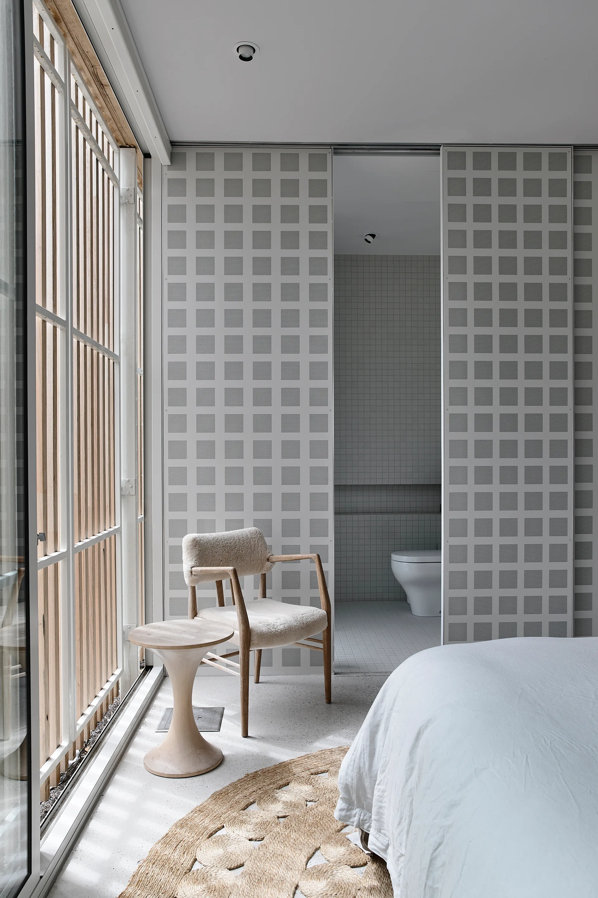 A minimalist bedroom features a sheepskin armchair and wood side table near a grey grid sliding door that opens to a tiled bathroom.