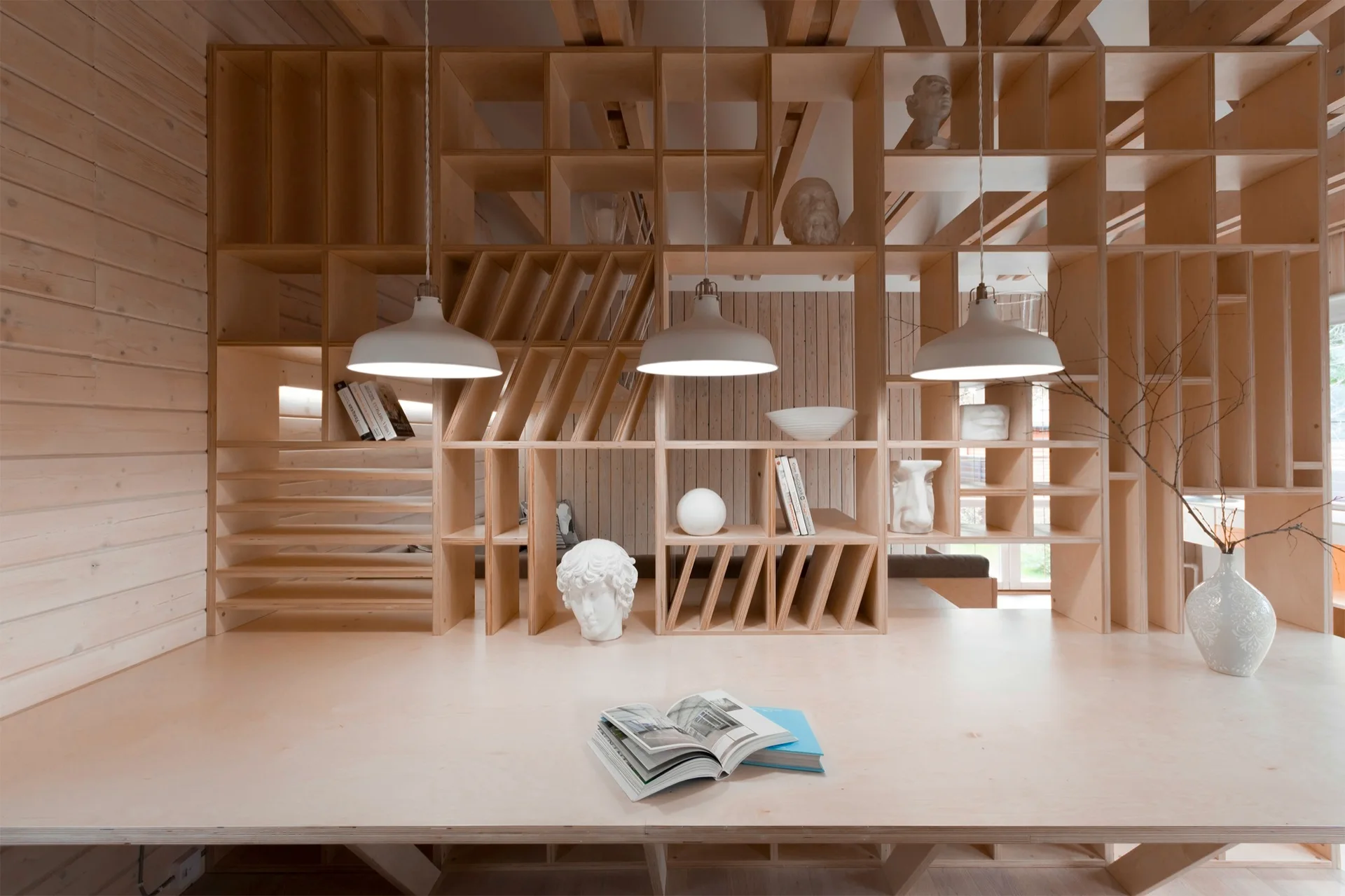 A spacious light plywood desk integrated with a custom modular grid shelving unit displaying white sculptures and three hanging white dome pendant lights.