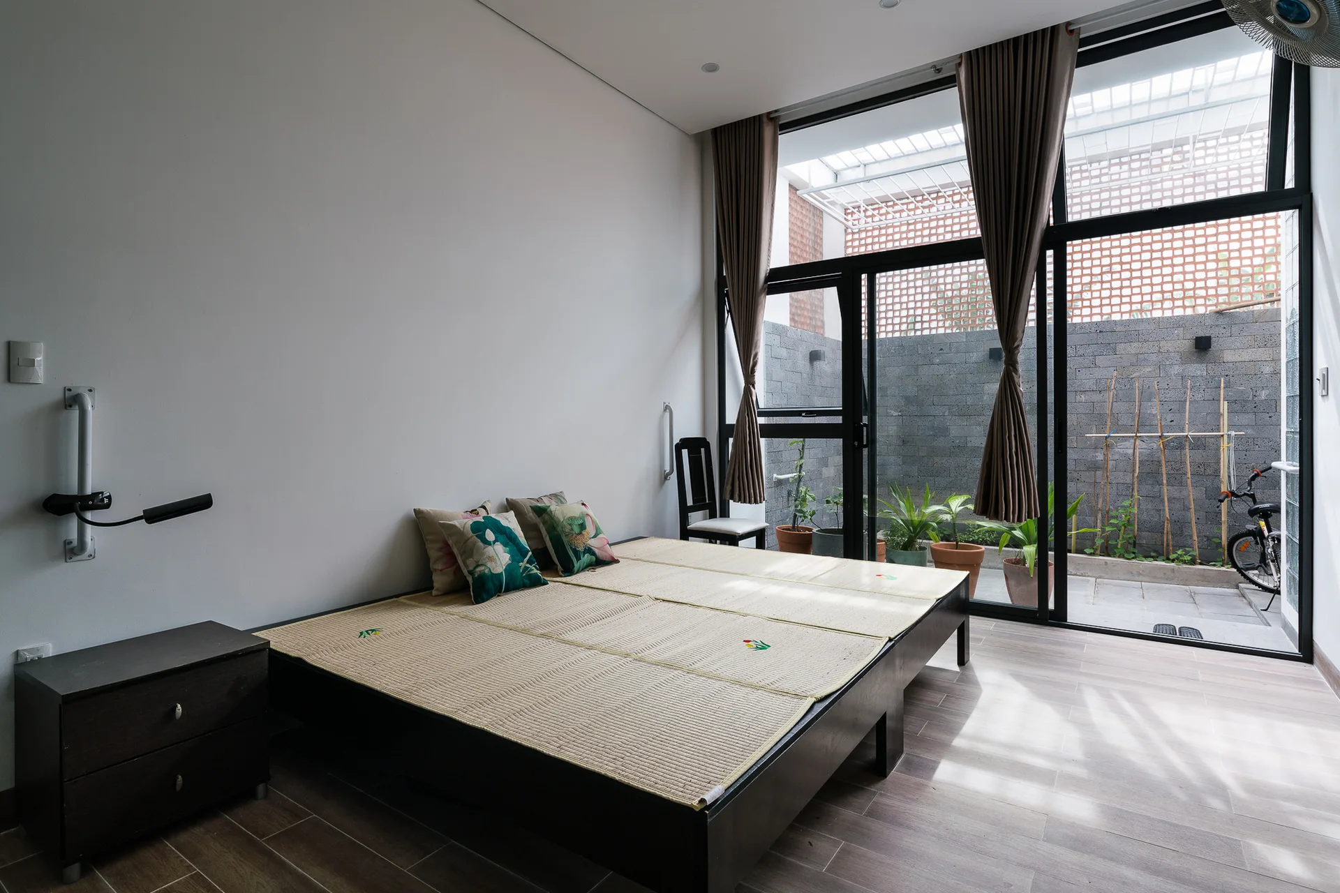 A minimalist bedroom featuring a low wooden platform bed with woven mats next to large glass doors opening to a small outdoor courtyard.