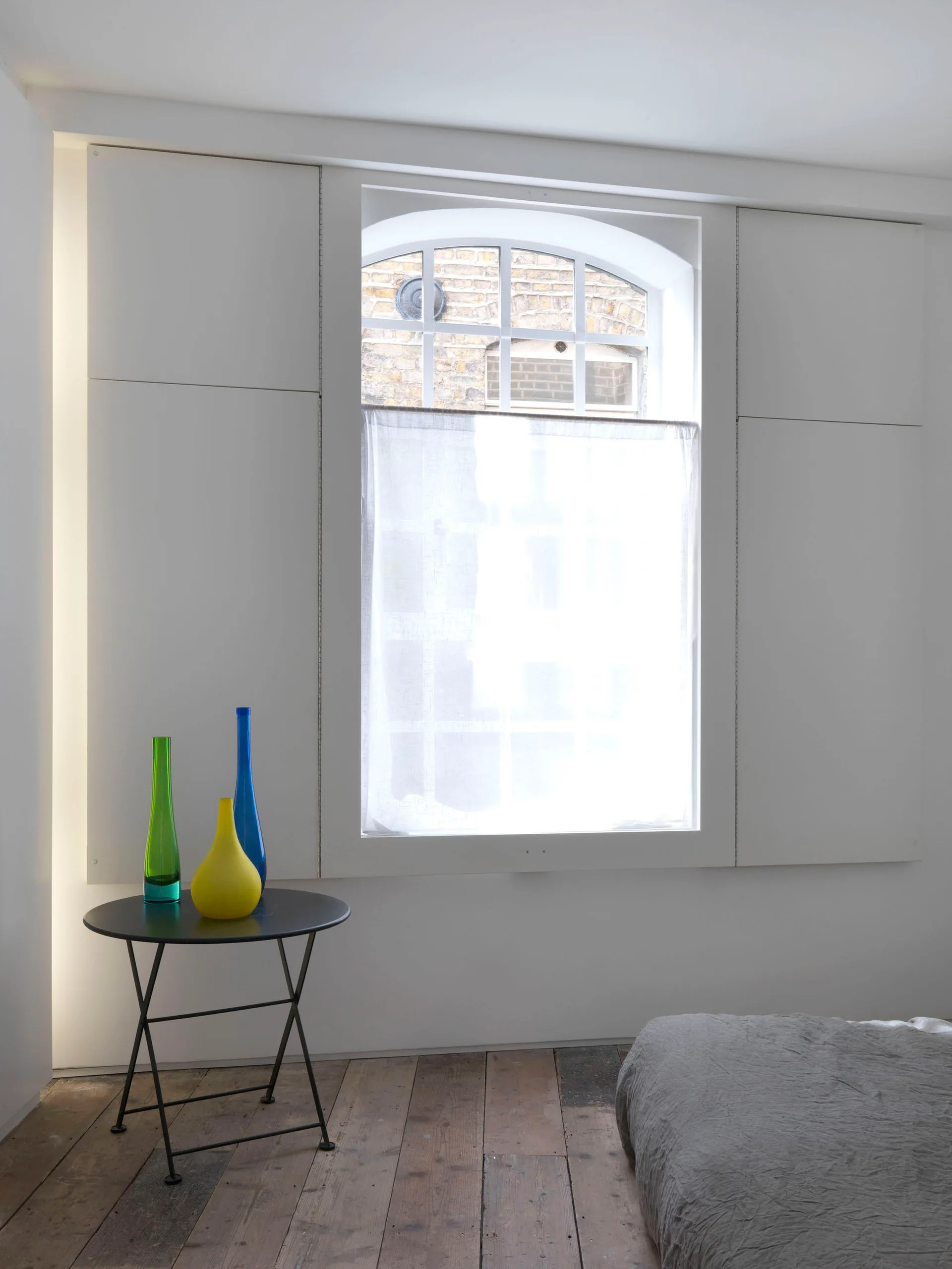 A minimalist bedroom features an arched window, a white paneled wall, and a small black side table topped with green, yellow, and blue glass vases.