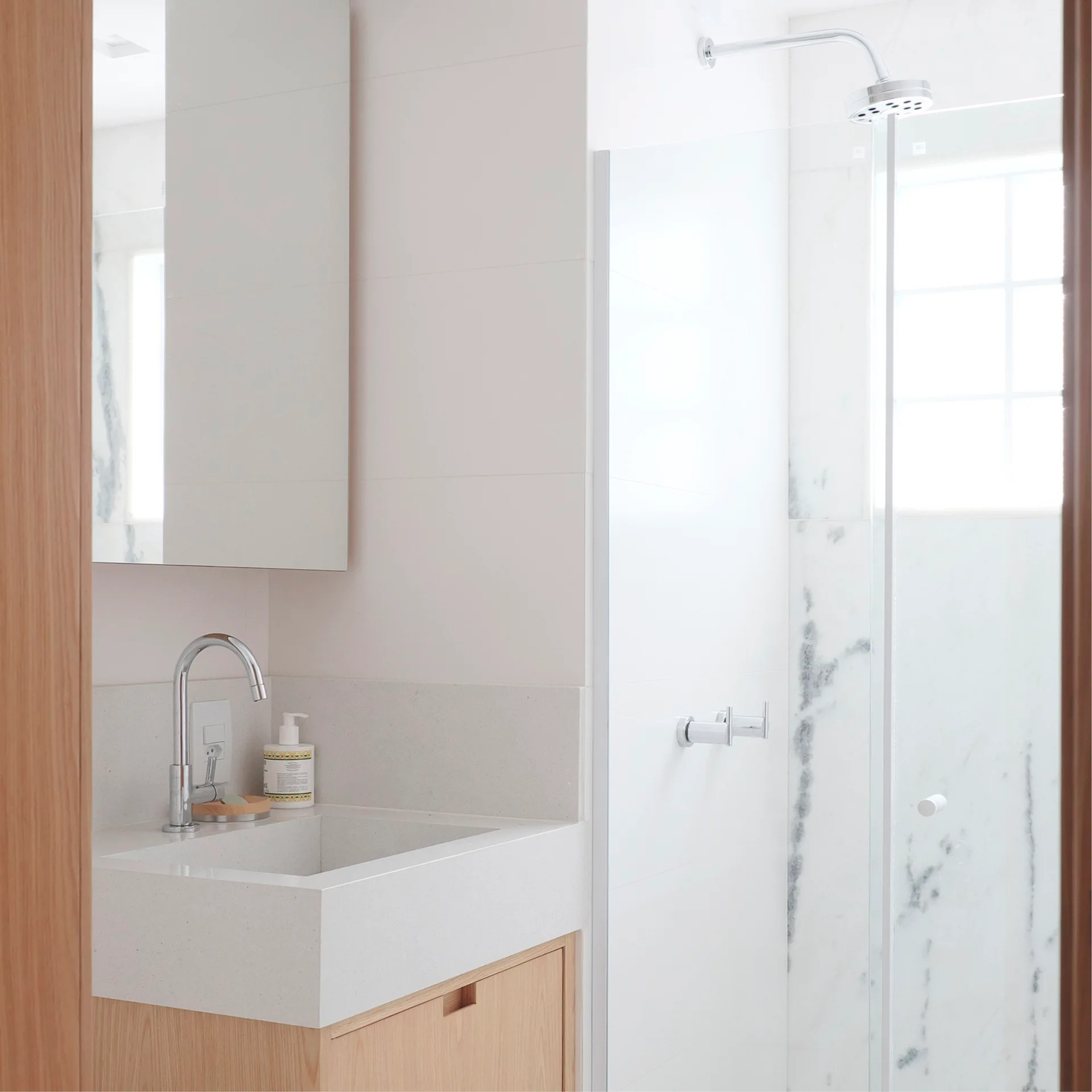 A modern bathroom features a light wood vanity with an integrated quartz sink, chrome faucet, and a glass-enclosed shower with white marble wall tiles.