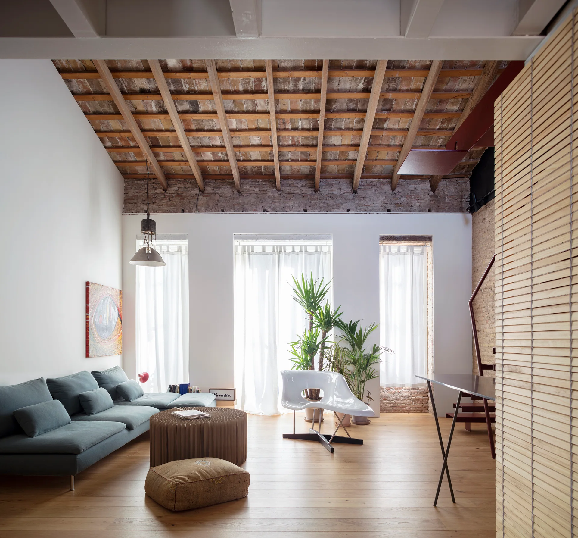 A loft living room features a teal sectional sofa, a white sculptural chair, and three windows beneath an exposed wood beam ceiling.