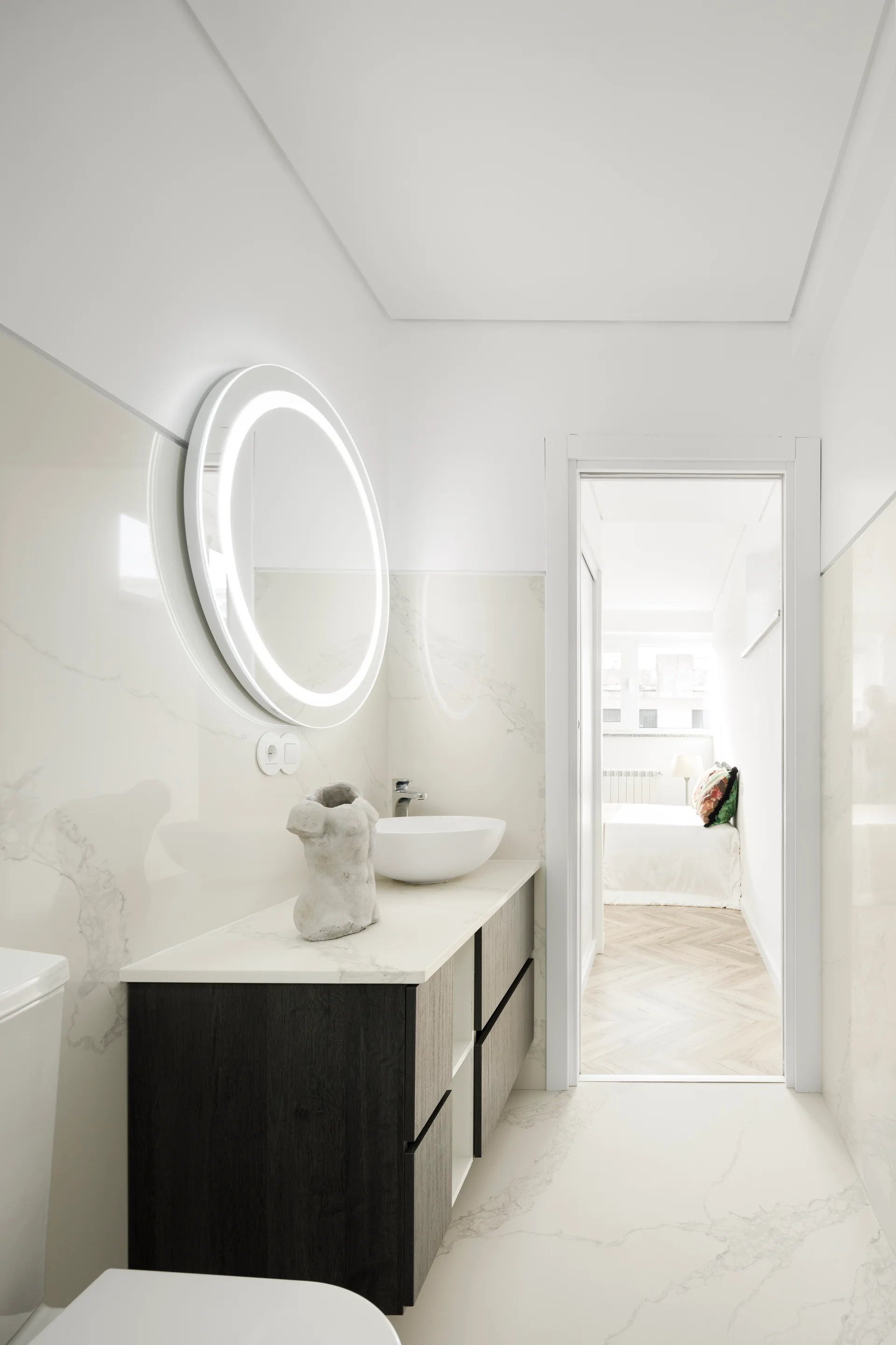 A modern bathroom features a dark wood floating vanity, a round backlit mirror, and marble-patterned wall tiles leading to an open bedroom doorway.