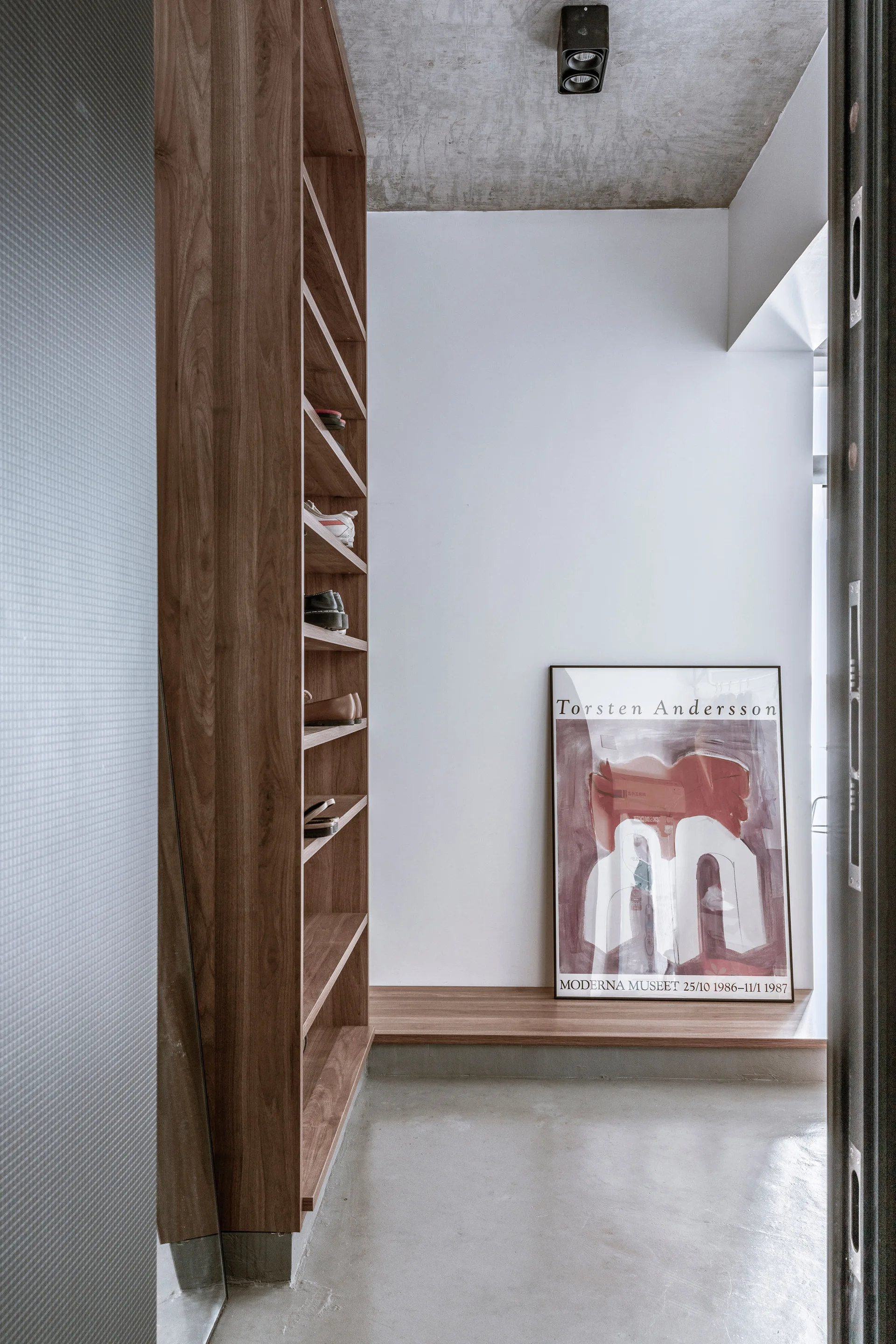 A minimalist entryway features a tall wooden shoe storage unit, a grey concrete floor and ceiling, and a framed art poster leaning on a low wood ledge.