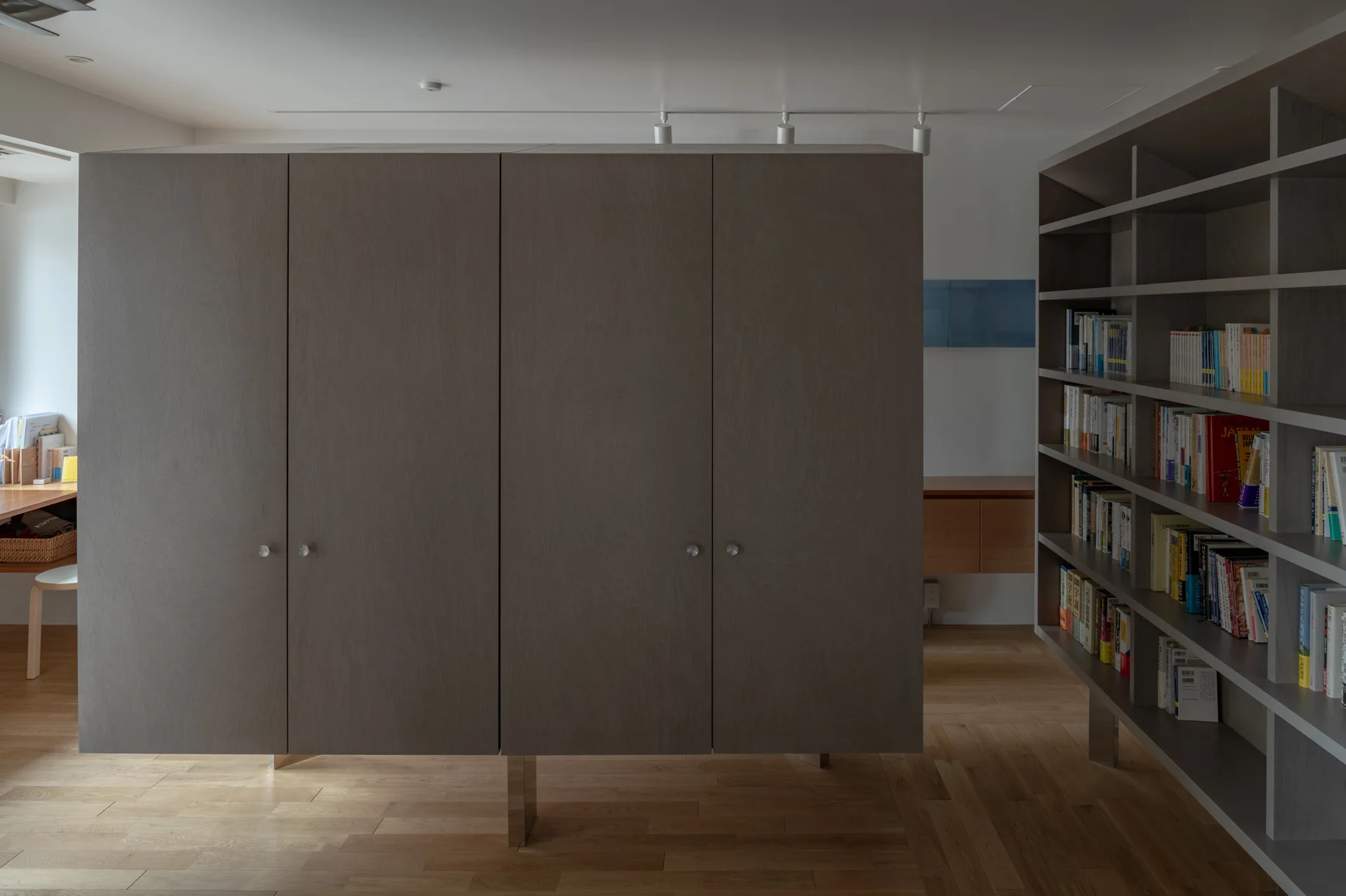 A large gray-brown wooden cabinet stands on metal legs in the center of a room with light wood floors next to a tall, filled open bookshelf.