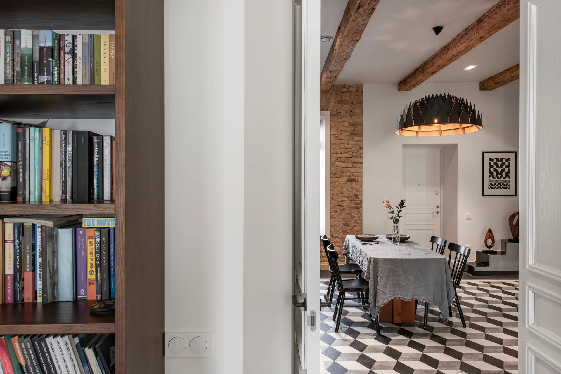 A dining room viewed through a doorway features checkered tile floors, rustic wood ceiling beams, a linen-covered table, and spindle chairs.