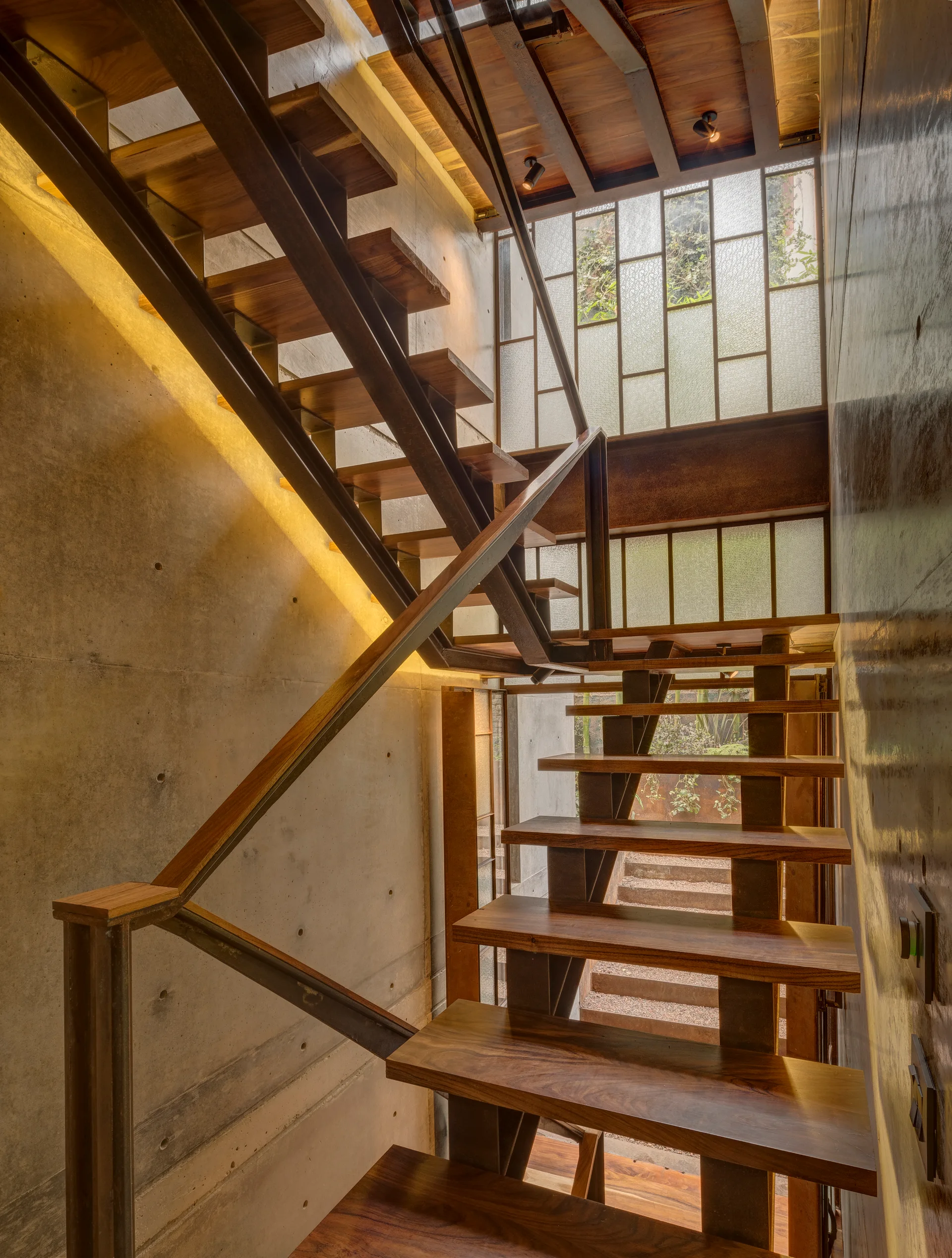 A floating industrial staircase features warm wood treads, a dark steel frame, exposed concrete walls, and a large patterned glass grid window.
