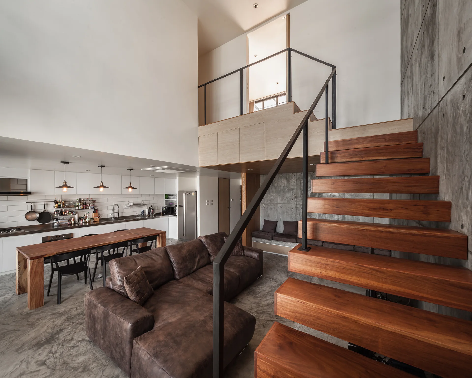 An industrial loft space features a prominent floating wooden staircase, a brown sectional sofa, and raw concrete walls.